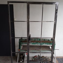 Artur Trawiński - Dwie metalowe ramy z podziałami, prawdopodobnie do drzwi lub okien, oparte o ścianę w warsztacie, widoczna zieleń maszyny do obróbki metalu i narzędzia.