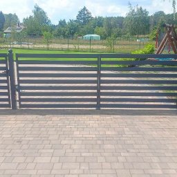 Artur Trawiński - Nowoczesna, pozioma, metalowa brama wjazdowa w kolorze grafitowym, z kulistymi zakończeniami słupków, na tle zielonego trawnika i drzew.