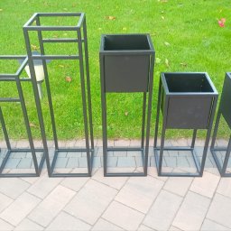 Artur Trawiński - Zestaw pięciu metalowych kwietników w różnych rozmiarach, ustawionych na brukowanym chodniku przed trawnikiem i zielenią.