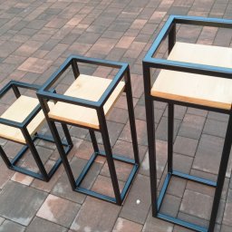 Artur Trawiński - Trzy metalowe kwietniki w stylu industrialnym z drewnianymi blatami, ustawione na kostce brukowej, różnej wysokości.