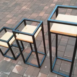 Trzy metalowe kwietniki w stylu industrialnym z drewnianymi blatami, ustawione na kostce brukowej, różnej wysokości.