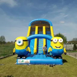 Niebiesko-żółty dmuchany zamek z motywem Minionków na zielonym trawniku, dziecko wchodzi po schodach na zjeżdżalnię, błękitne niebo w tle.