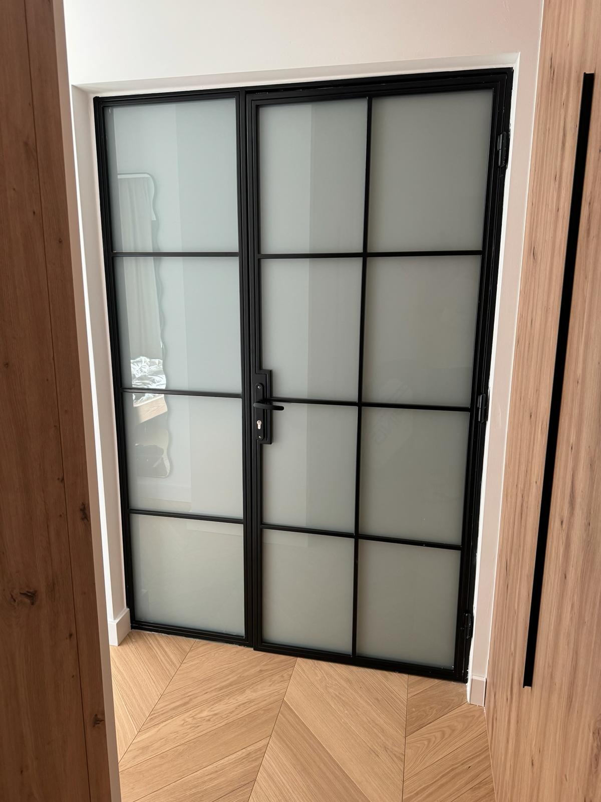 Nowoczesne, dwuskrzydłowe drzwi wewnętrzne z czarną ramą i matowym szkłem, widoczne na tle parkietu ułożonego w jodełkę.