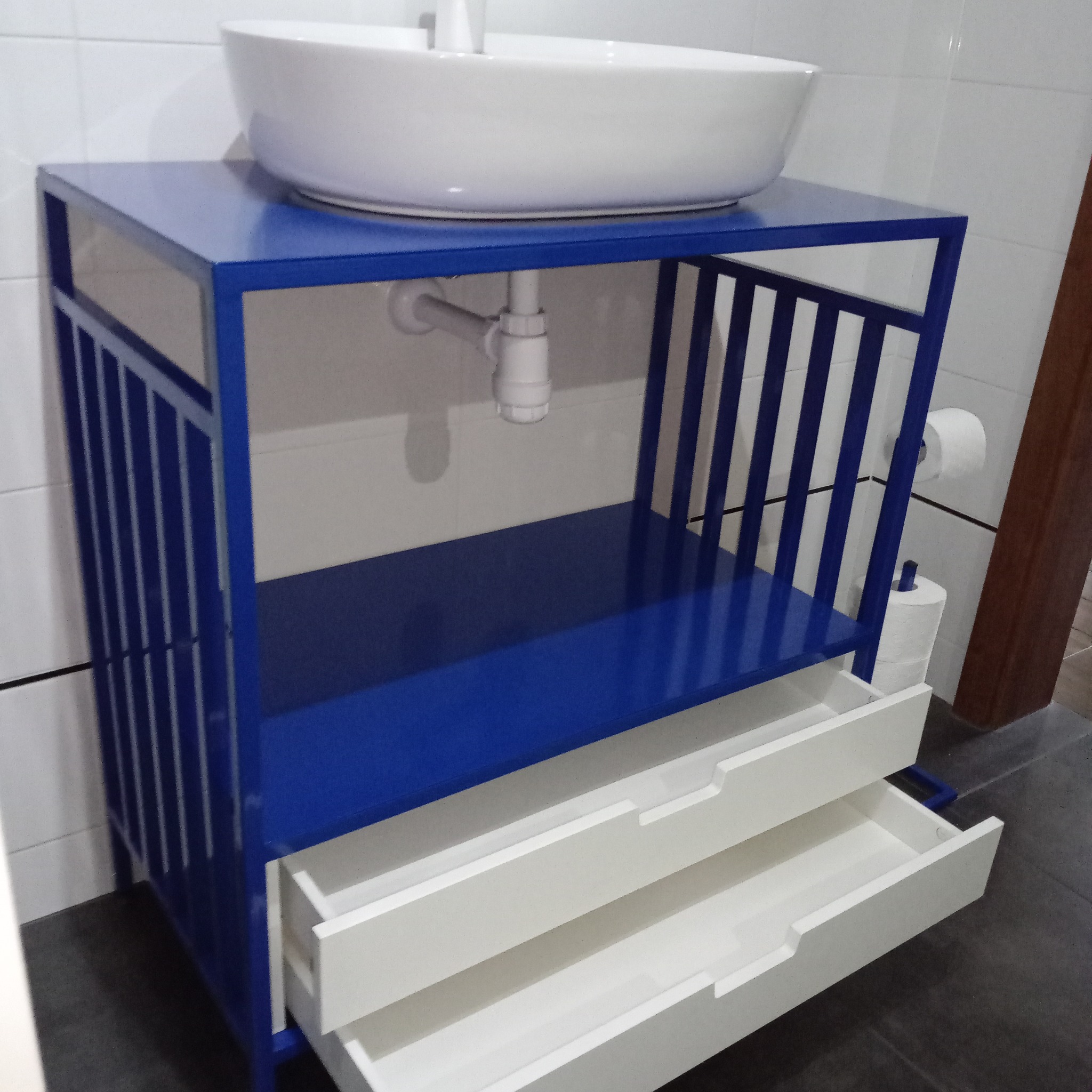 Niebieska metalowa szafka łazienkowa z otwartymi białymi szufladami i okrągłą umywalką nablatową, widoczne rury odpływowe i toaleta w tle.