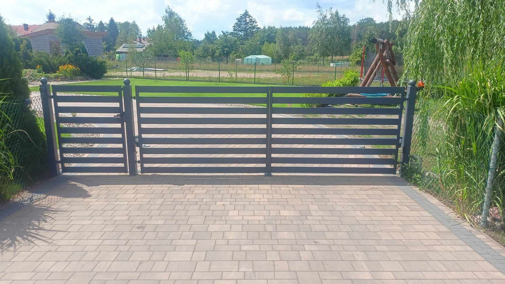 Nowoczesna, pozioma, metalowa brama wjazdowa w kolorze grafitowym, z kulistymi zakończeniami słupków, na tle zielonego trawnika i drzew.