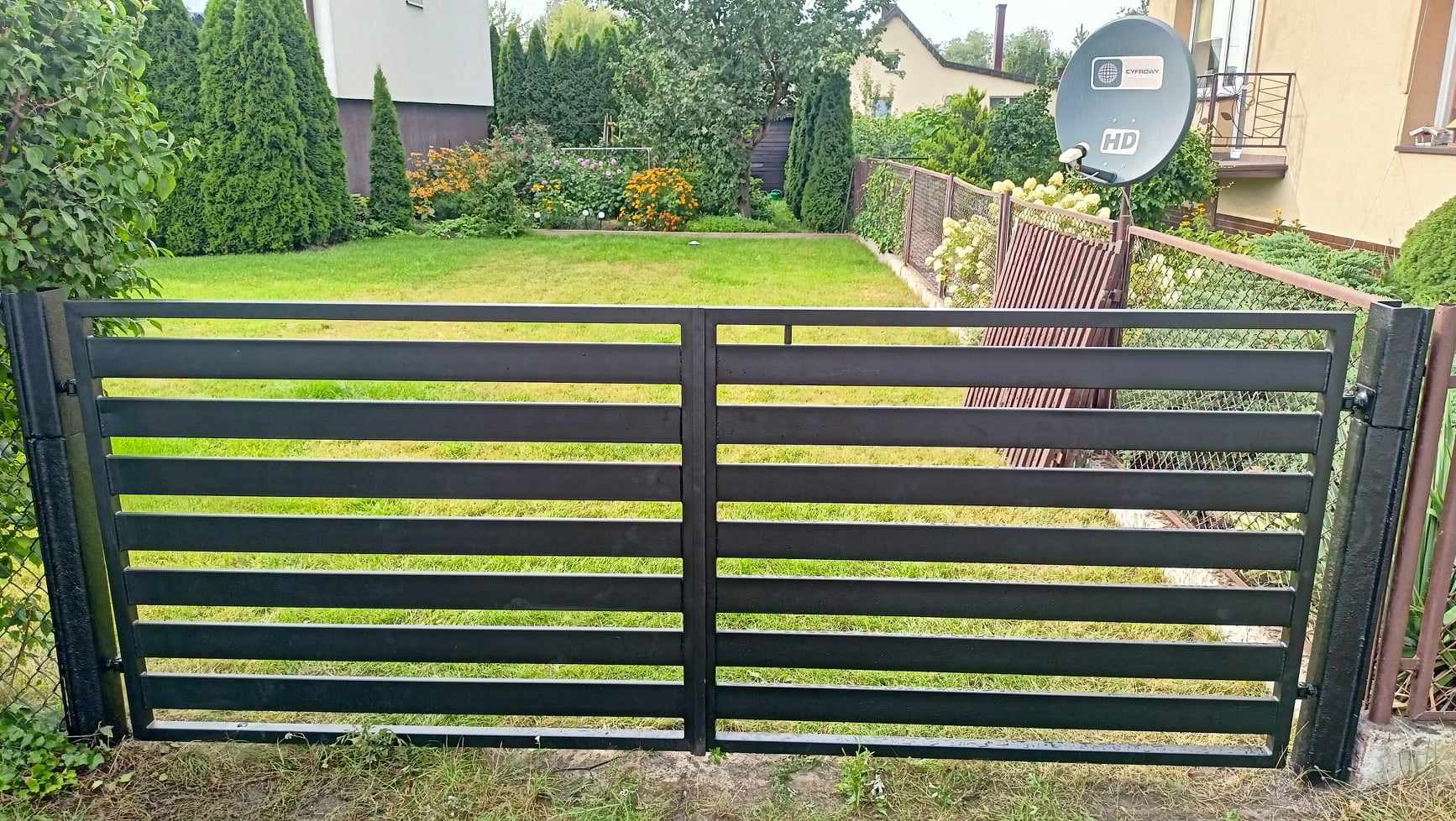 Nowoczesna, czarna, metalowa brama dwuskrzydłowa z poziomymi panelami na tle zielonego trawnika i ogrodu.
