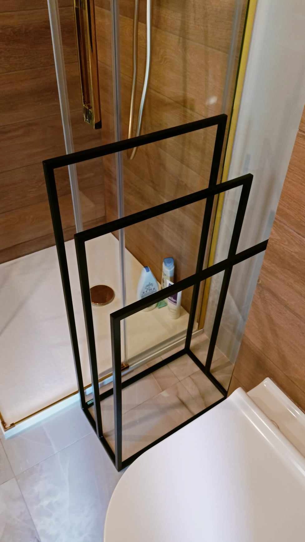 Czarny, metalowy stojak na ręczniki w łazience z kabiną prysznicową i drewnopodobnymi płytkami na ścianach.