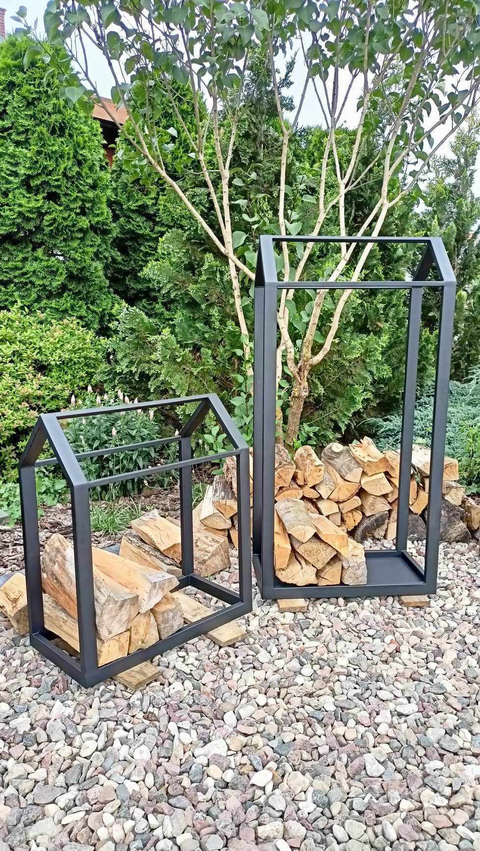 Dwa metalowe stojaki na drewno kominkowe, jeden w kształcie domku, drugi wyższy z podstawą, ustawione na żwirowym podłożu w ogrodzie.