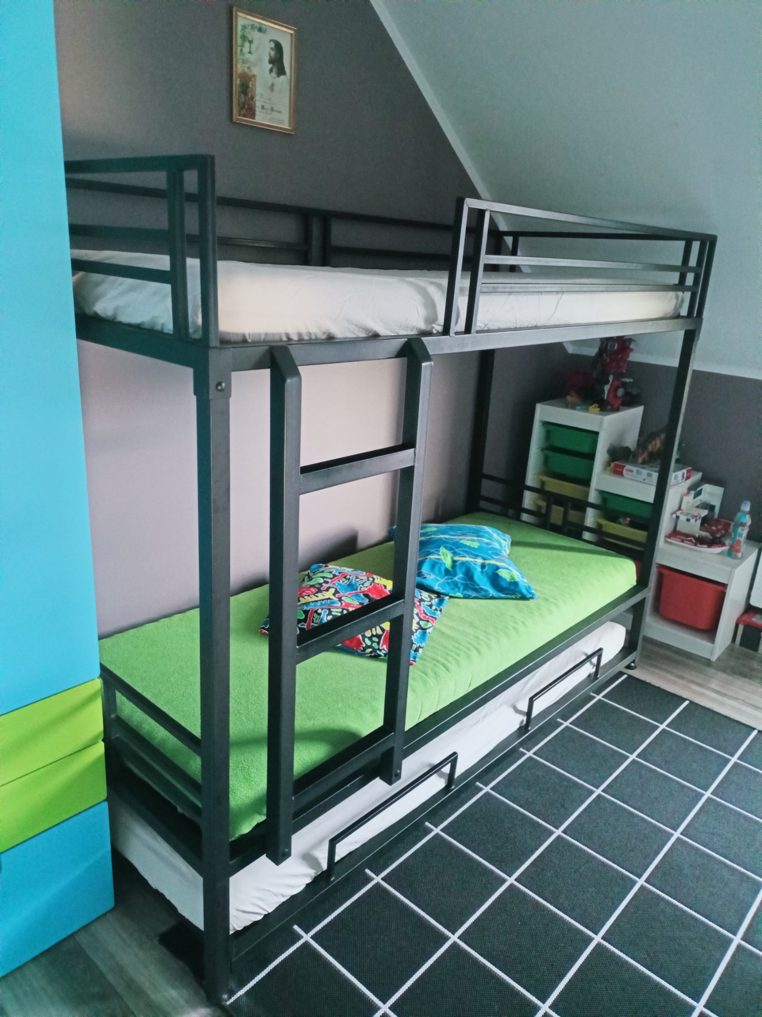 Metalowa rama łóżka piętrowego z drabinką, zielonym materacem na dole i białym na górze, na tle szarej ściany i kolorowych szafek.