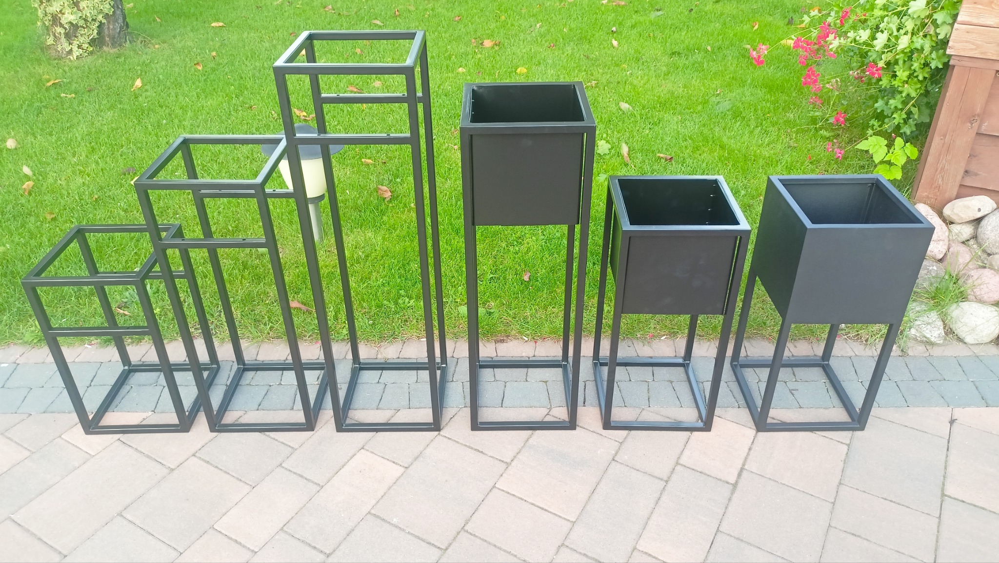 Zestaw pięciu metalowych kwietników w różnych rozmiarach, ustawionych na brukowanym chodniku przed trawnikiem i zielenią.
