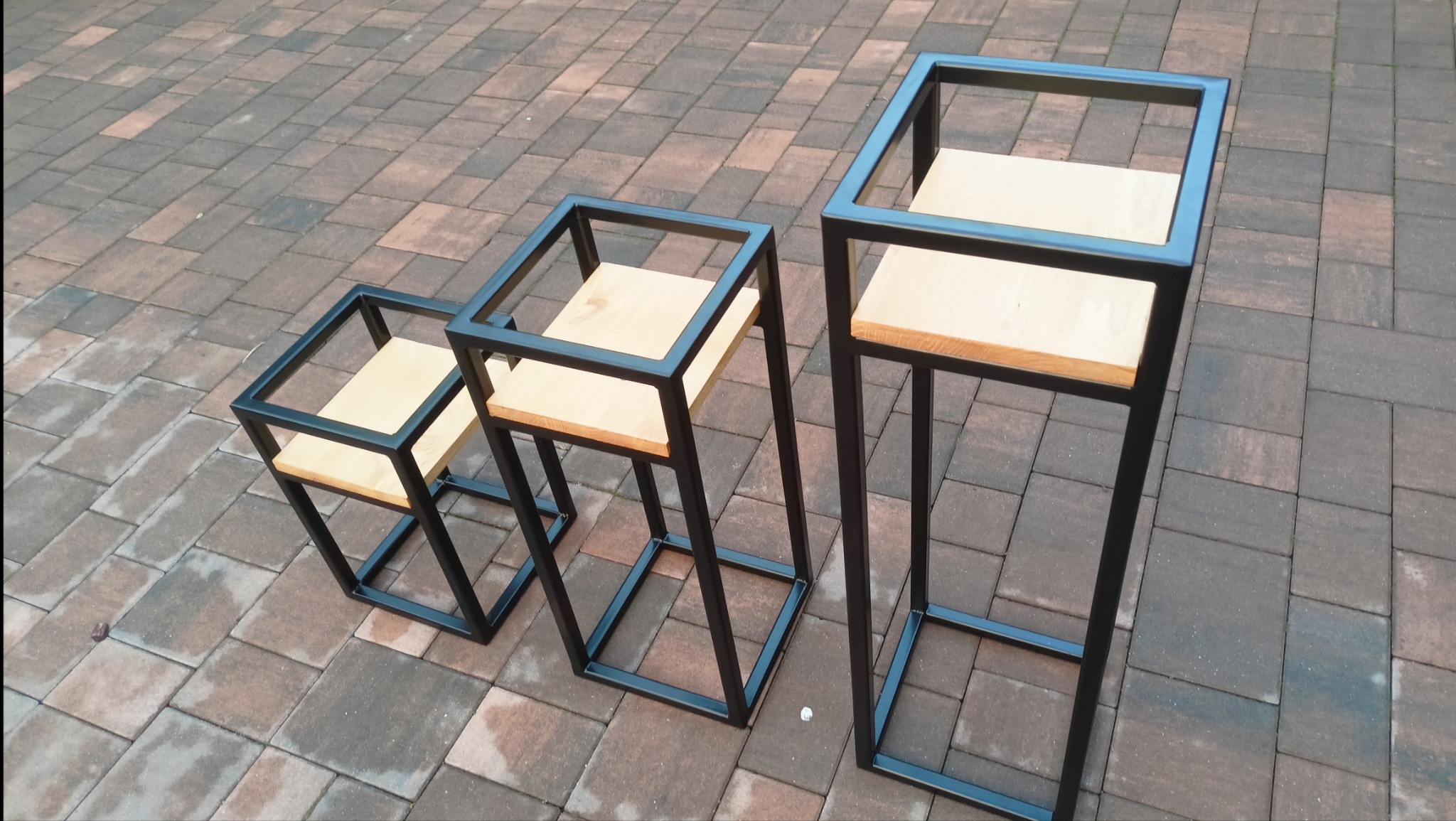 Trzy metalowe kwietniki w stylu industrialnym z drewnianymi blatami, ustawione na kostce brukowej, różnej wysokości.