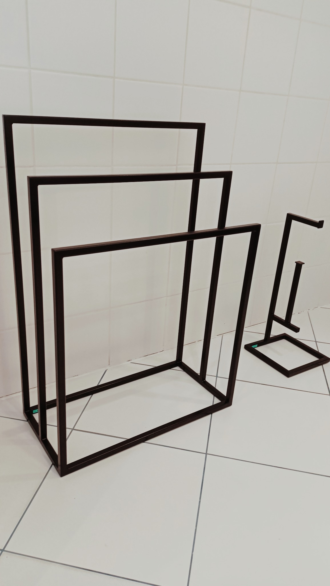 Metalowe stojaki na ręczniki i papier toaletowy w minimalistycznym stylu, pomalowane na ciemny kolor, ustawione na białych płytkach podłogowych i ściennych.