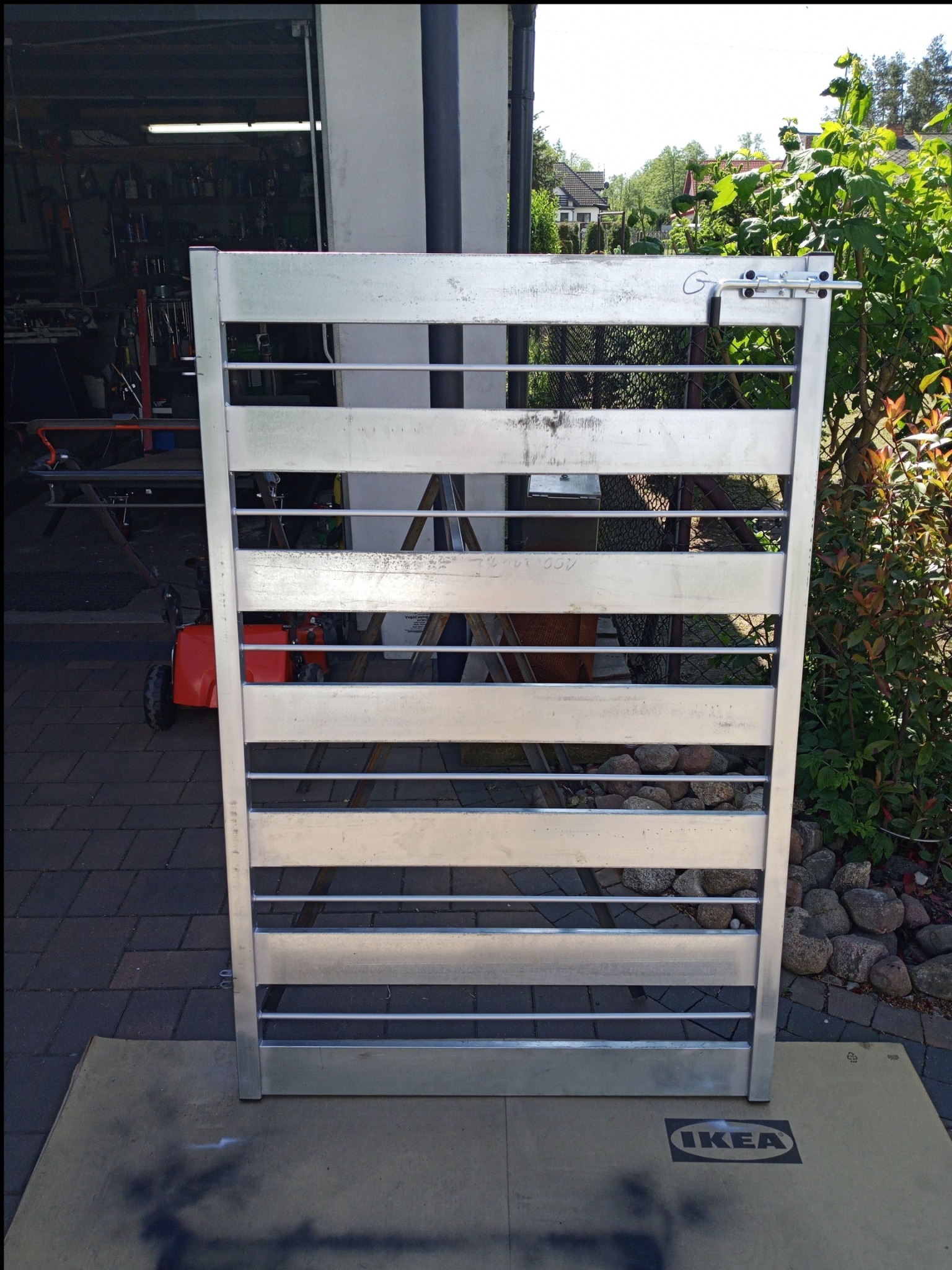 Surowa, aluminiowa konstrukcja bramki z poziomymi szczeblami, z widocznym zamkiem, oparta na kartonie z logo IKEA, na tle warsztatu i zieleni ogrodowej.