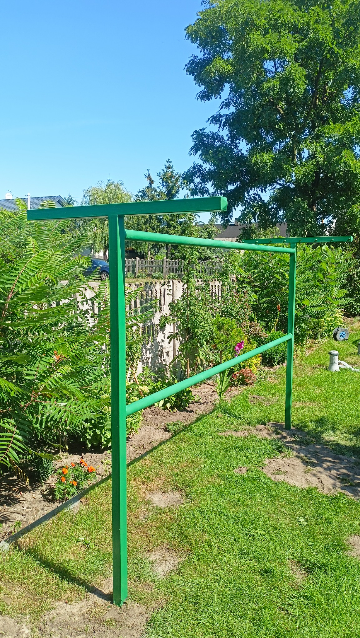 Zielona metalowa konstrukcja w ogrodzie, prawdopodobnie pergola w trakcie montażu, widoczne elementy mocujące, bujna roślinność w tle.