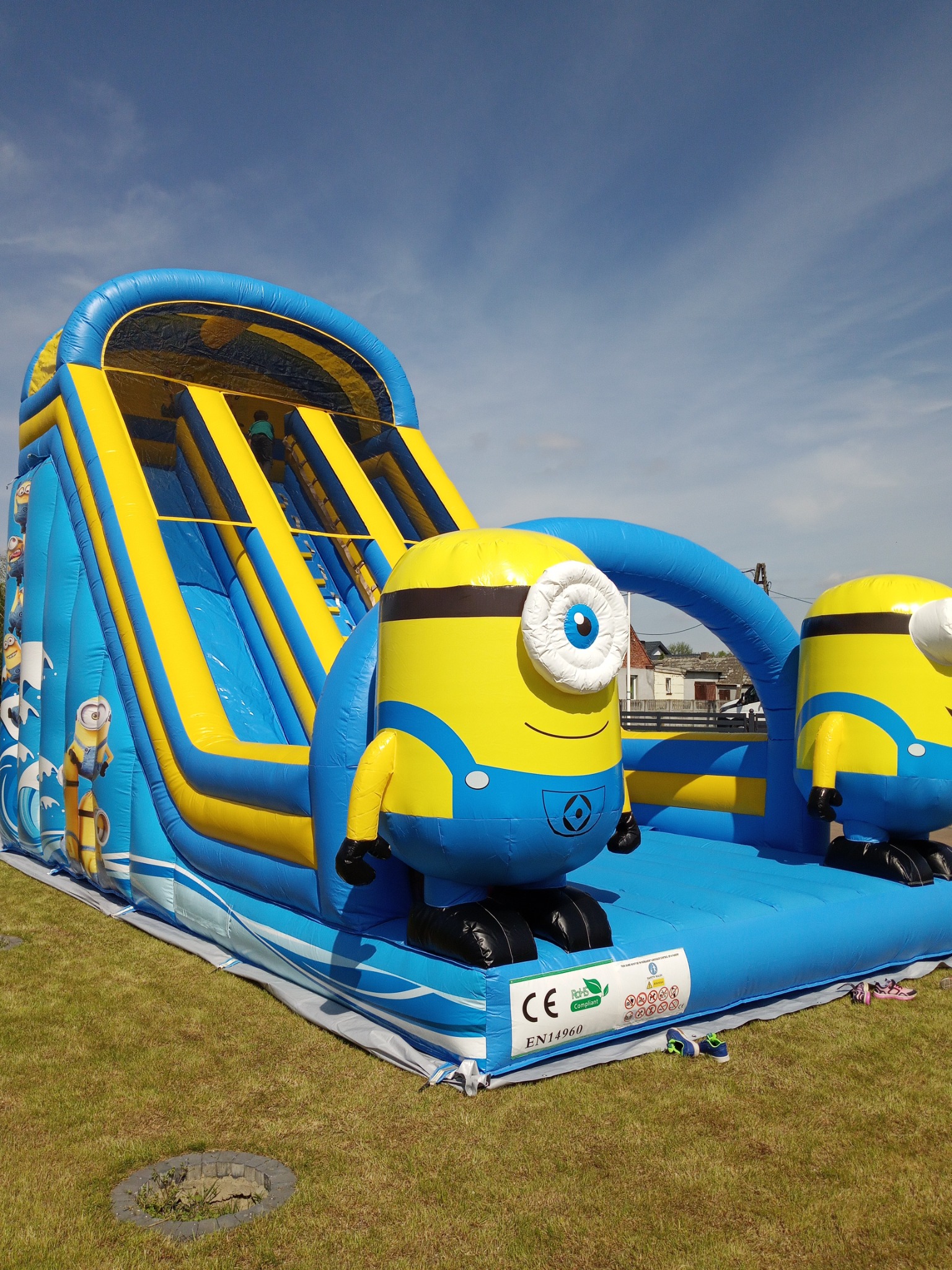 Niski kąt ujęcia na niebiesko-żółty, dmuchany zamek ze zjeżdżalnią z motywem Minionków na trawniku; widoczne dzieci na szczycie zjeżdżalni.