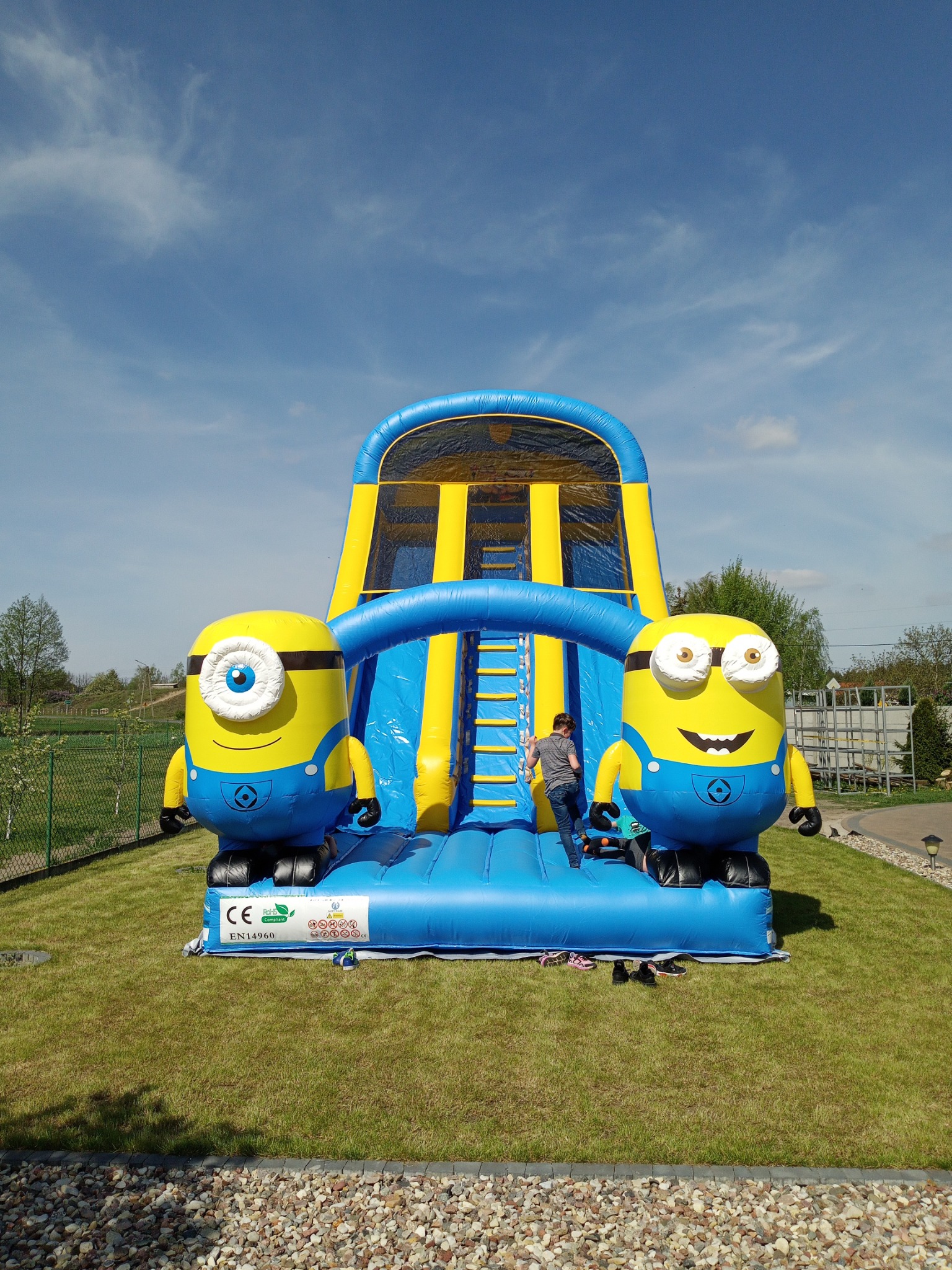 Niebiesko-żółty dmuchany zamek z motywem Minionków na zielonym trawniku, dziecko wchodzi po schodach na zjeżdżalnię, błękitne niebo w tle.