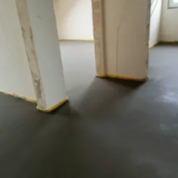 Wylana, ciemnoszara posadzka w trakcie wiązania, zabezpieczona żółtą taśmą przy ścianach i ościeżnicach, w tle widoczne surowe ściany i instalacja elektryczna.