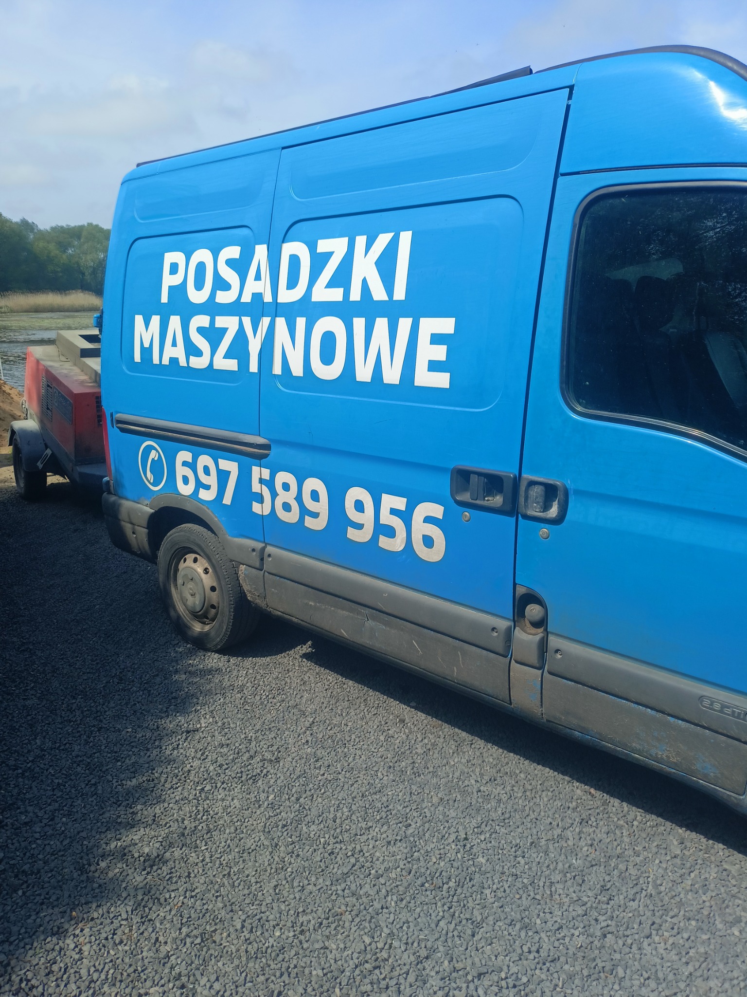 Niebieski samochód dostawczy z białym napisem 'POSADZKI MASZYNOWE' i numerem telefonu, stojący na żwirowym podłożu, z przyczepą z maszyną do posadzek w tle.