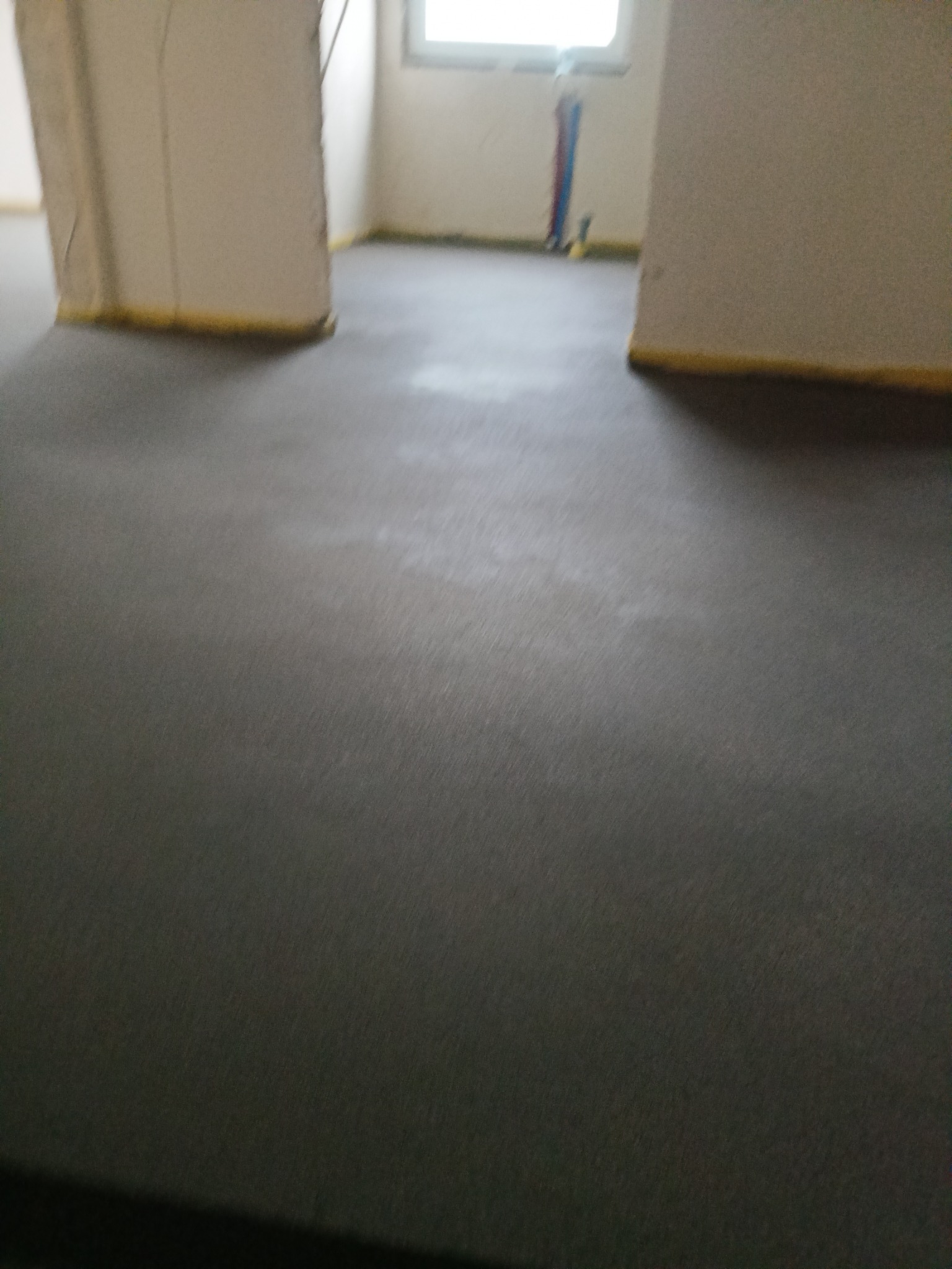 Wylana, świeża, szara posadzka w pomieszczeniu z widocznymi rurami instalacji na ścianie w tle i żółtą taśmą zabezpieczającą przy ścianach.