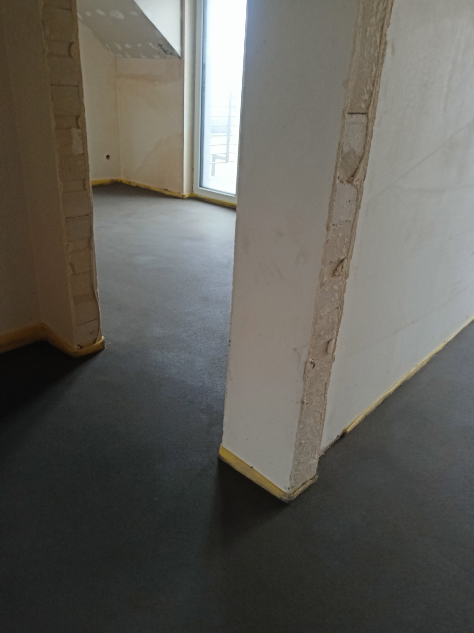 Świeżo wylana, ciemna posadzka w surowym wnętrzu, zabezpieczona żółtą taśmą przy ścianach i filarach.