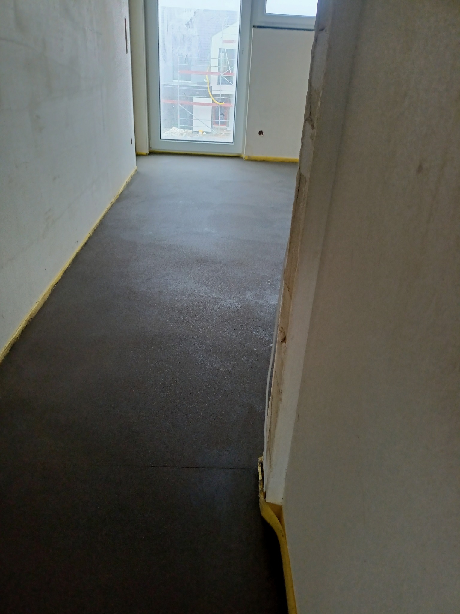 Świeżo wylana, ciemnoszara posadzka w surowym pomieszczeniu z oknem, zabezpieczona żółtą taśmą przy ścianach.