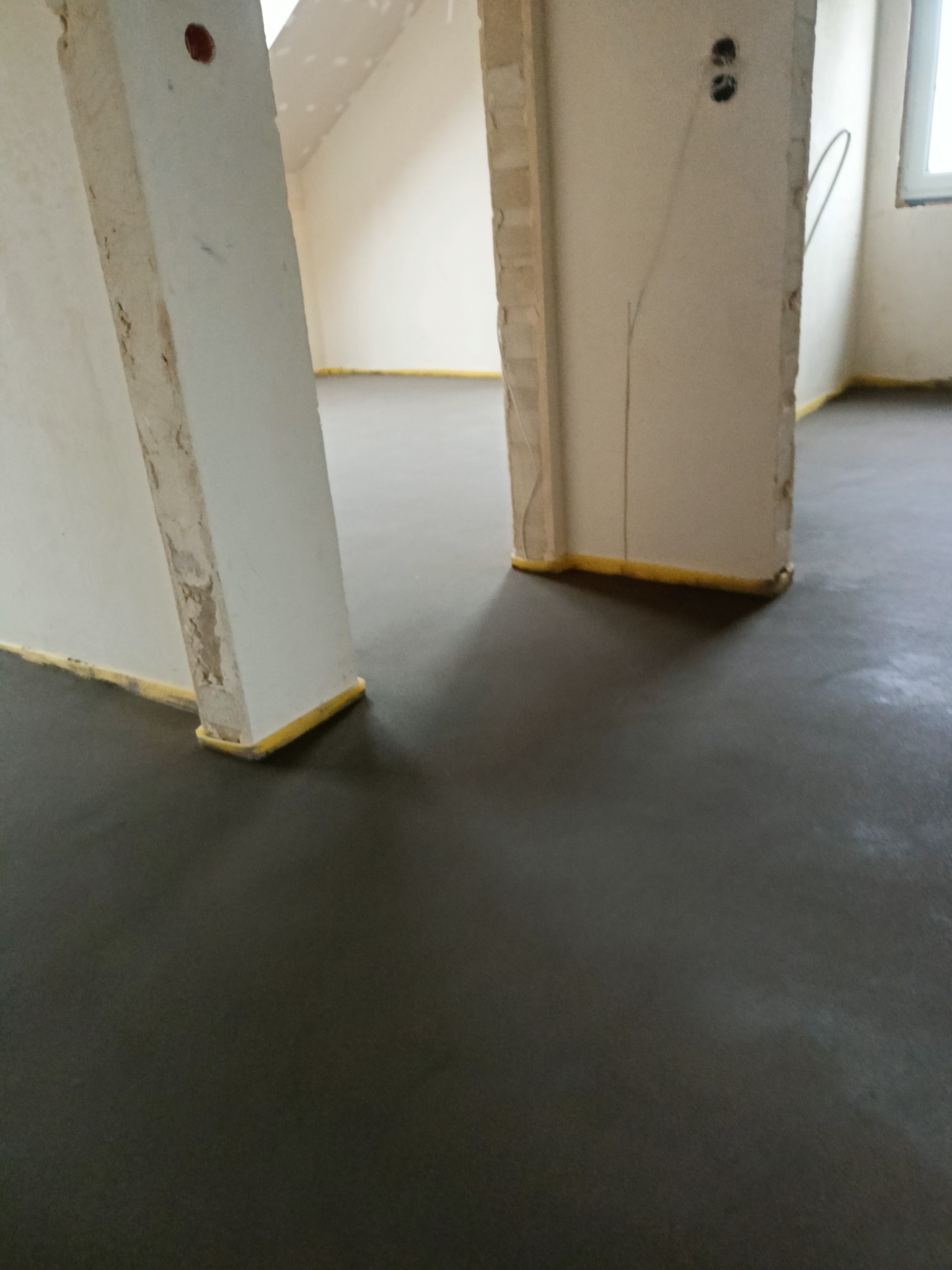 Wylana, ciemnoszara posadzka w trakcie wiązania, zabezpieczona żółtą taśmą przy ścianach i ościeżnicach, w tle widoczne surowe ściany i instalacja elektryczna.