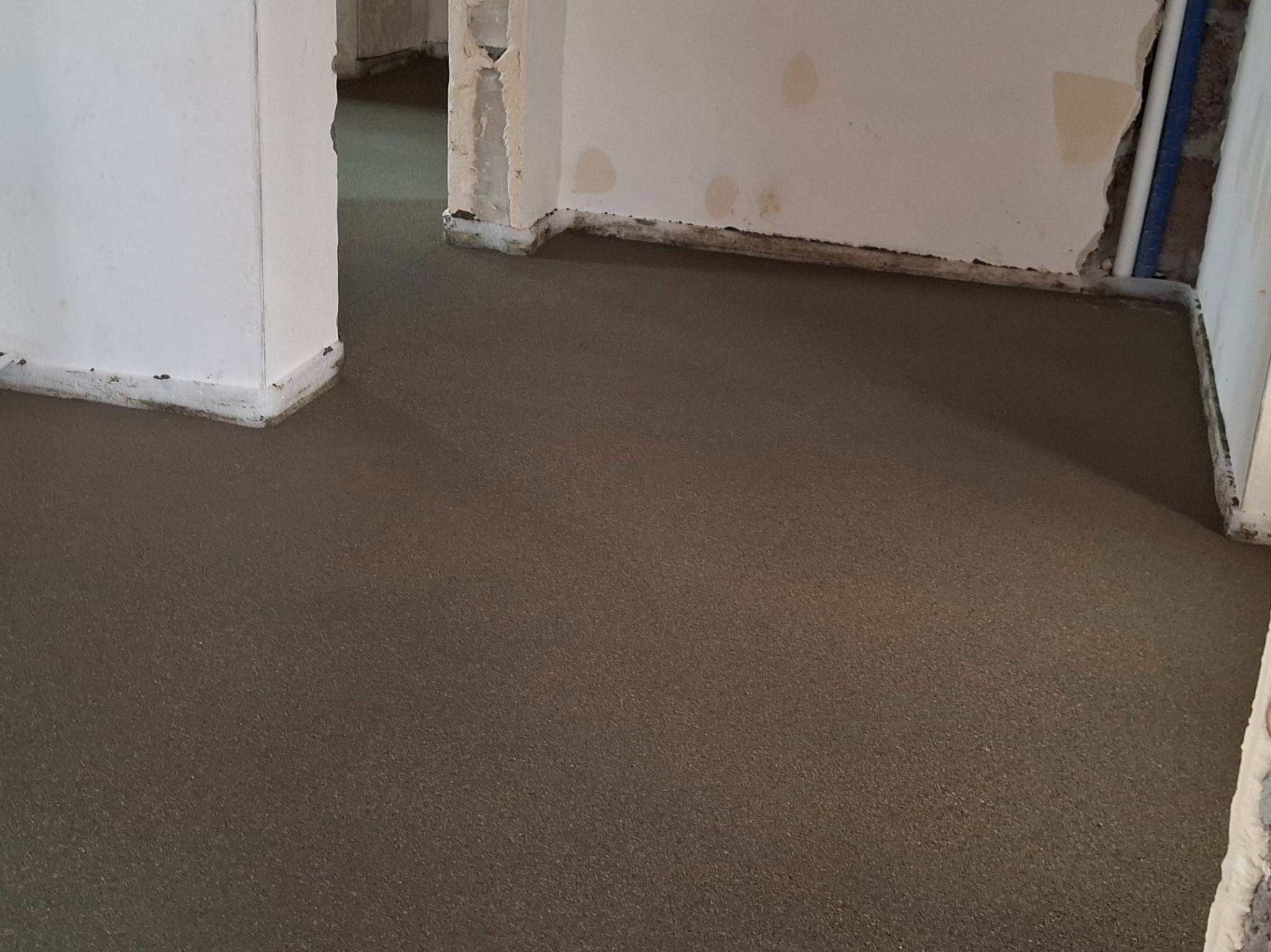 Świeżo wylana, gładka posadzka w surowym wnętrzu. Widoczne fragmenty ścian z resztkami tynku i wystające rury. Minimalistyczny kadr na posadzkę.