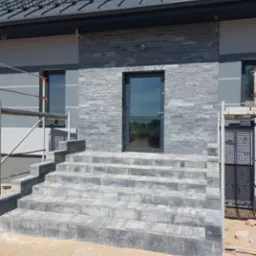 Elewacja domu z szarej cegły i schodami z kostki. Widoczne rusztowania, okna z ciemnymi ramami i ciemne drzwi wejściowe. Nowoczesny design.