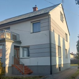 Eugeniusz Harasim - Dwukondygnacyjny dom z szarą elewacją i elementami dekoracyjnymi, balkon z metalową balustradą, schody wejściowe wyłożone terakotą, w słoneczny dzień.