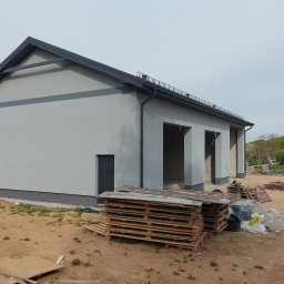 Eugeniusz Harasim - Szary budynek z ciemnymi elementami wykończeniowymi, widok z zewnątrz. Palety na pierwszym planie, teren wokół w trakcie prac budowlanych.