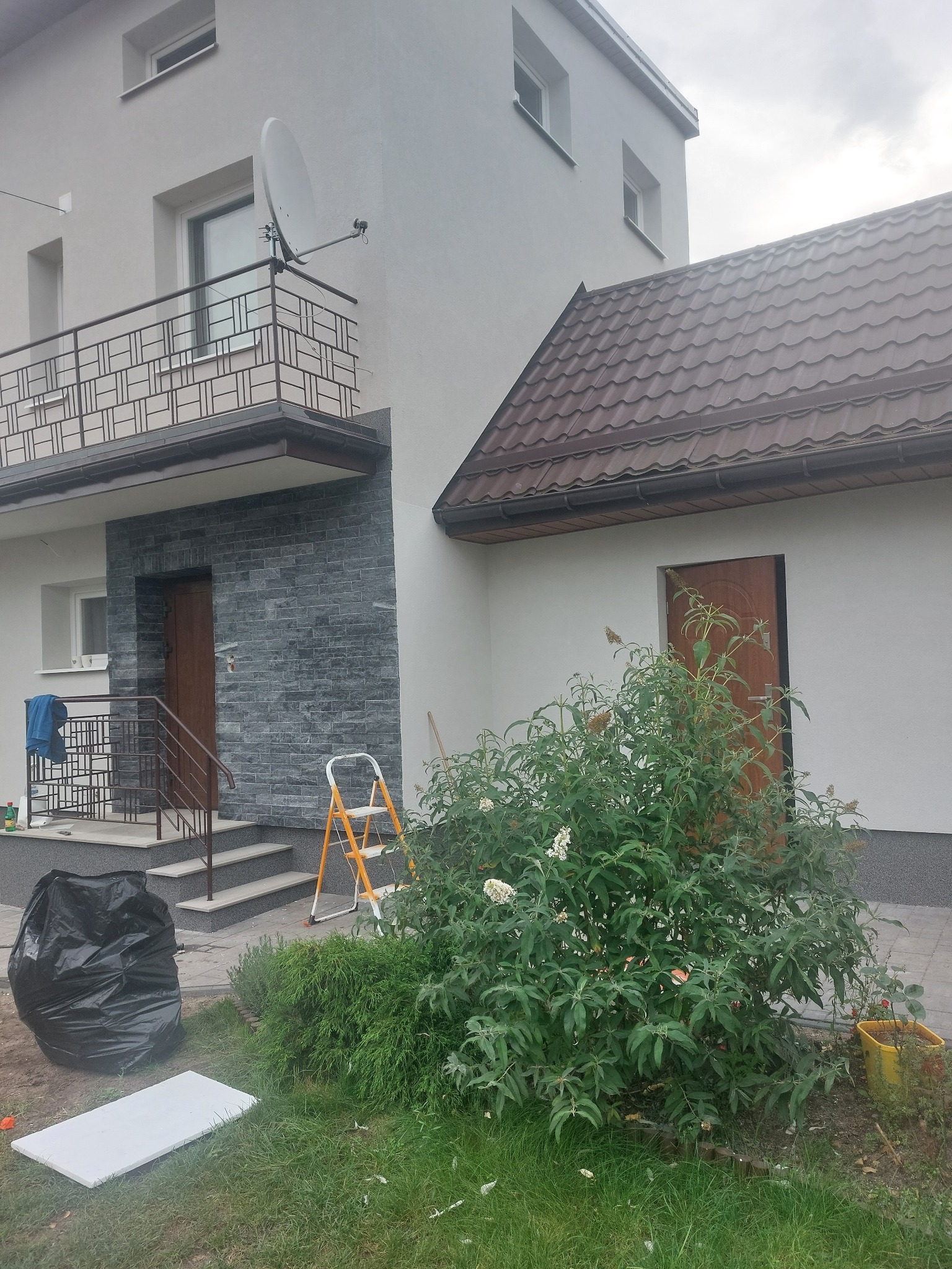 Wykończona elewacja domu z elementami dekoracyjnymi z kamienia i nową stolarką okienną. Widoczna drabina i worek na odpady sugerują prace wykończeniowe.