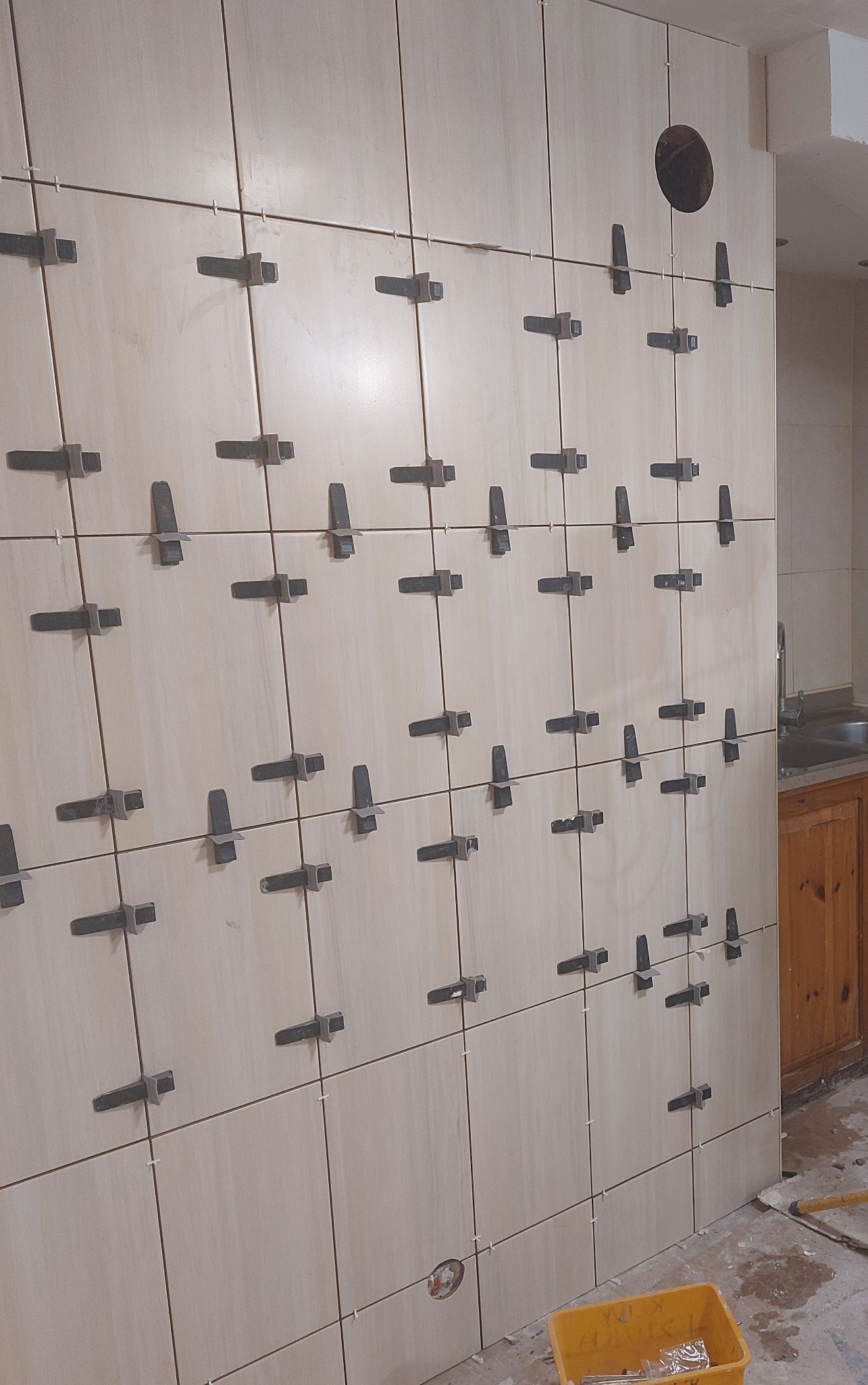 Jasne płytki ścienne w trakcie montażu, z użyciem klipsów poziomujących. Widoczne fugi i otwór w ścianie. Fragment szafki i podłogi z resztkami materiałów budowlanych.