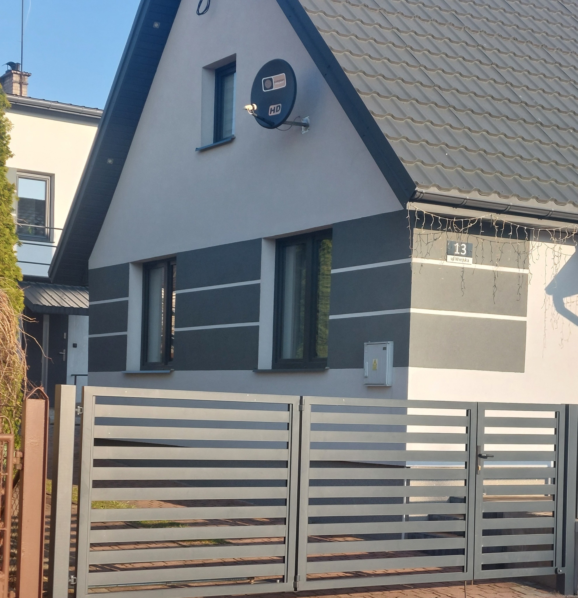 Dom jednorodzinny z szarą elewacją w poziome pasy, widoczny zza nowoczesnego, metalowego ogrodzenia. Na ścianie numer domu i antena satelitarna.