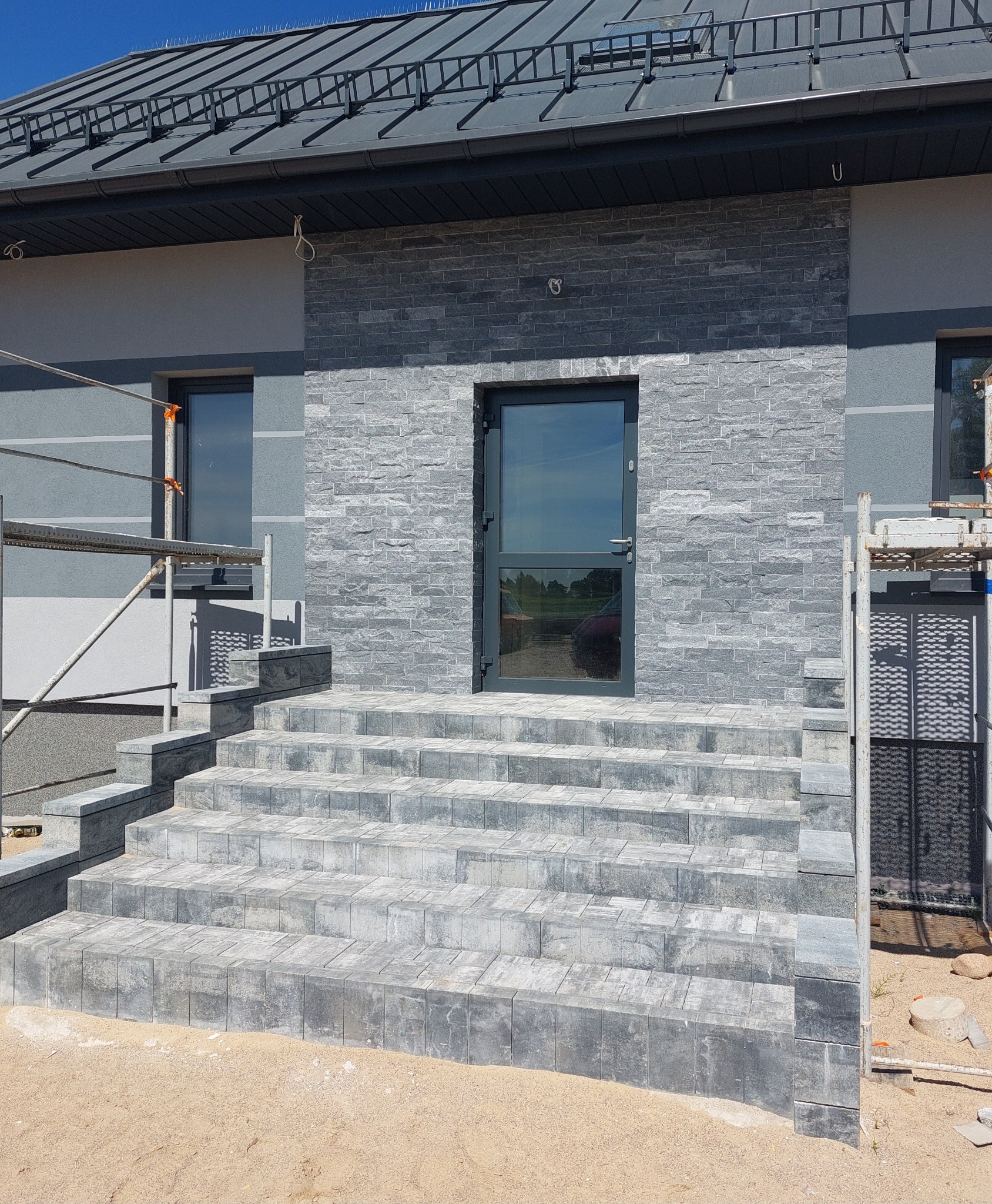 Elewacja domu z szarej cegły i schodami z kostki. Widoczne rusztowania, okna z ciemnymi ramami i ciemne drzwi wejściowe. Nowoczesny design.