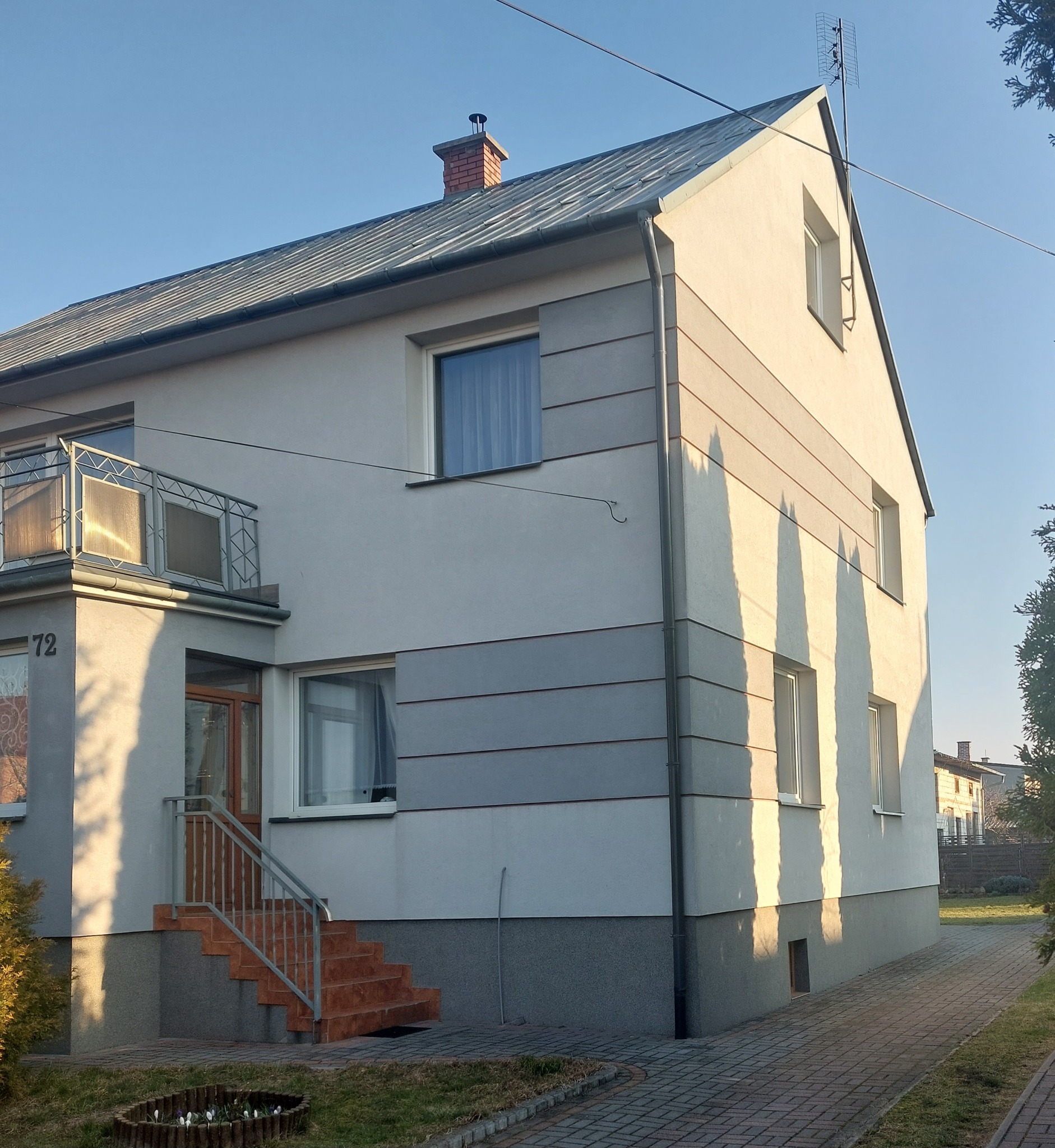 Dwukondygnacyjny dom z szarą elewacją i elementami dekoracyjnymi, balkon z metalową balustradą, schody wejściowe wyłożone terakotą, w słoneczny dzień.