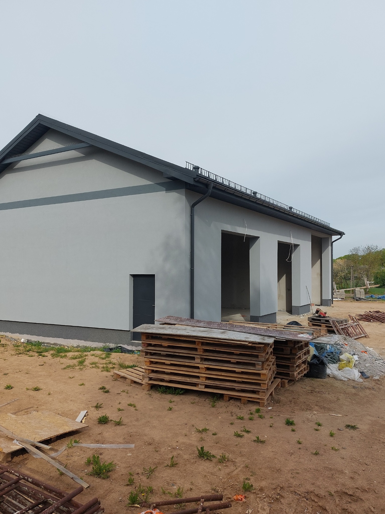 Szary budynek z ciemnymi elementami wykończeniowymi, widok z zewnątrz. Palety na pierwszym planie, teren wokół w trakcie prac budowlanych.