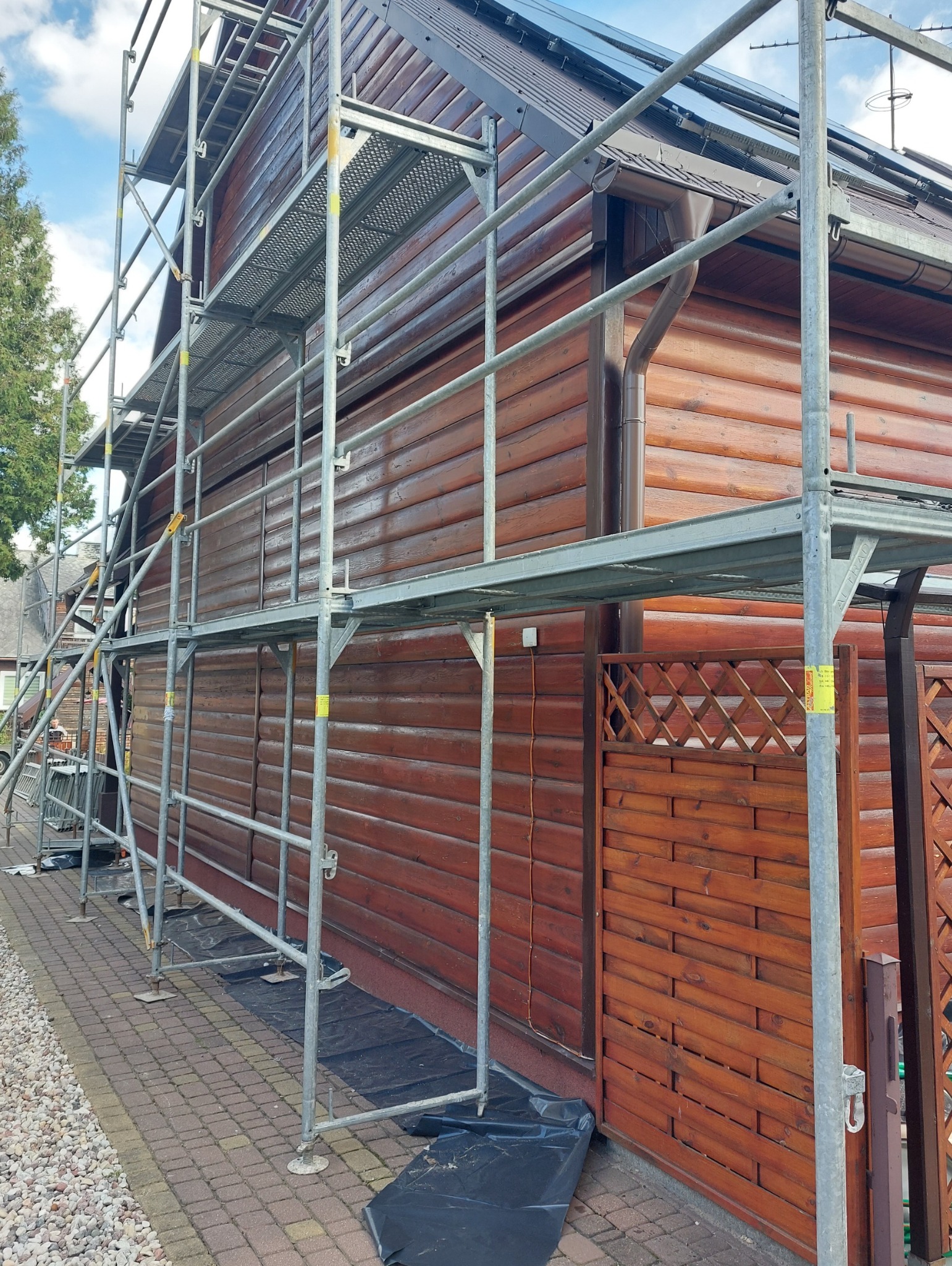 Drewniana elewacja domu jednorodzinnego w trakcie prac konserwacyjnych, widoczna z poziomu rusztowania. Świeżo polakierowane deski elewacyjne.