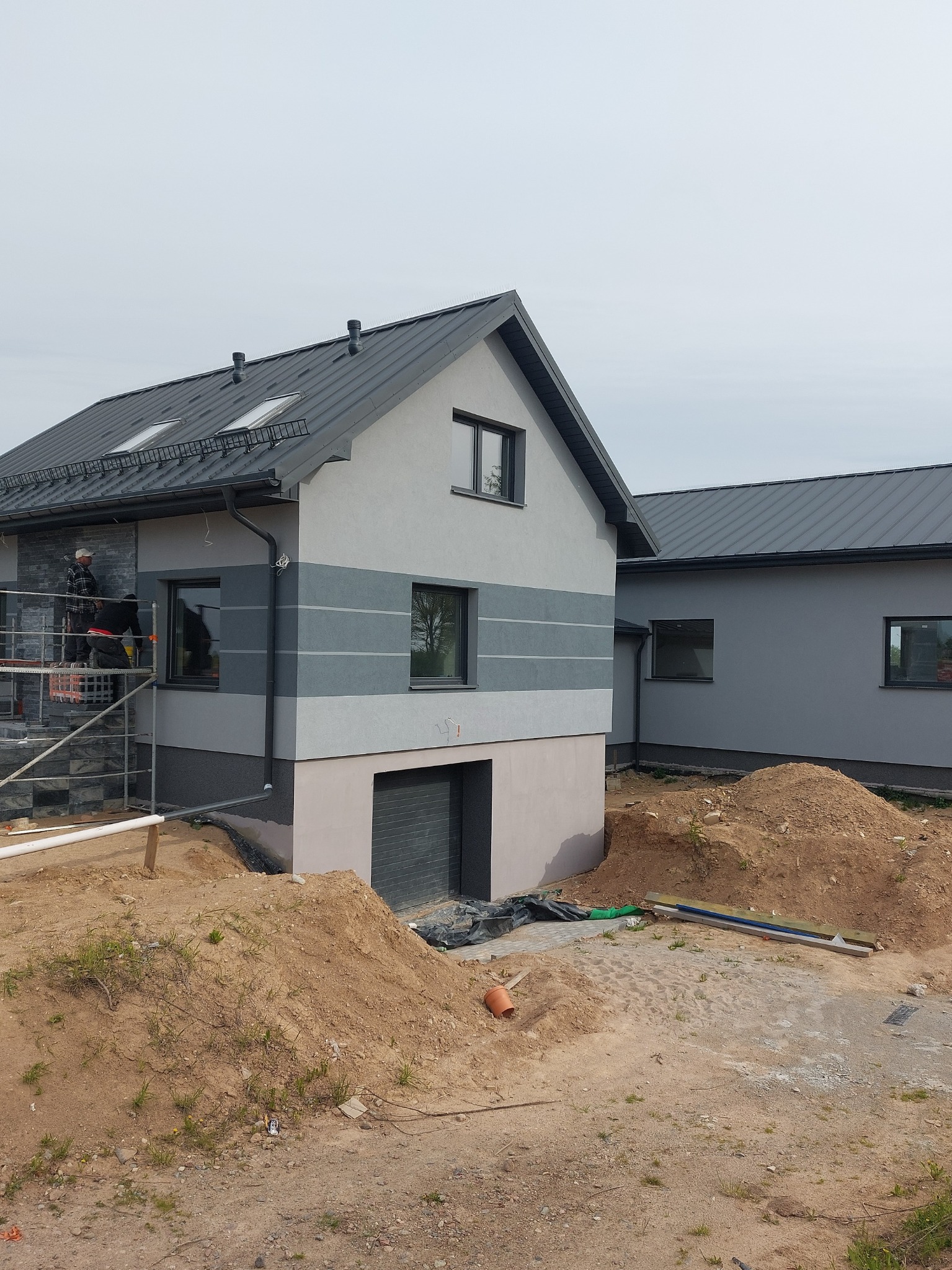 Dwukondygnacyjny dom z szarą elewacją w paski. Na rusztowaniu pracownicy montują kamień elewacyjny. Widoczne hałdy ziemi i materiały budowlane.