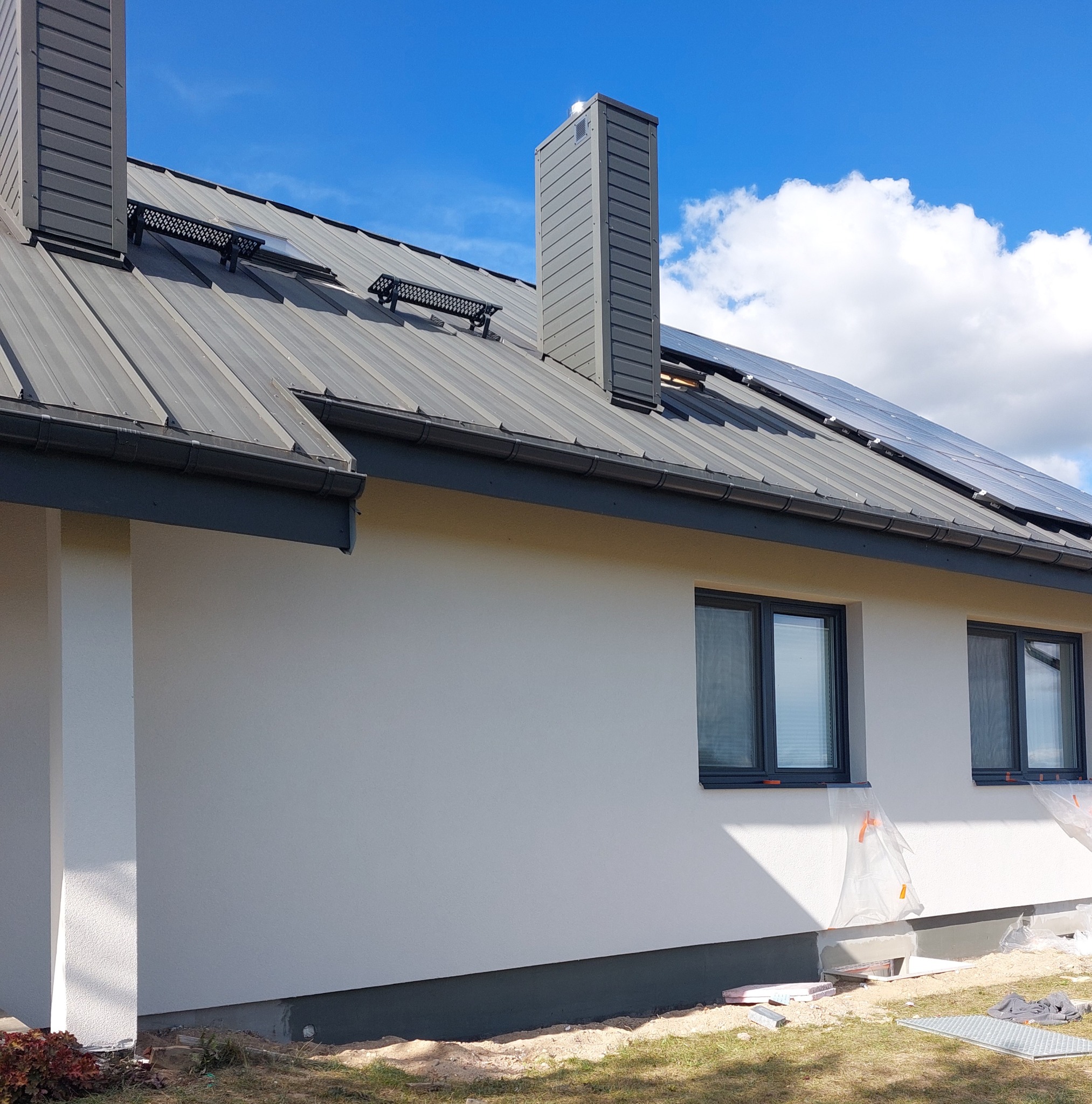 Elewacja domu jednorodzinnego po tynkowaniu, z widocznymi oknami w grafitowych ramach, metalowym dachem z panelami solarnymi i kominem w kolorze dachu.