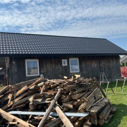DACH-MAT Mateusz Russu - Drewniany budynek z nowym, czarnym dachem z blachodachówki, na pierwszym planie sterta drewna i rusztowanie, w tle błękitne niebo z chmurami.