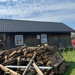 Drewniany budynek z nowym, czarnym dachem z blachodachówki, na pierwszym planie sterta drewna i rusztowanie, w tle błękitne niebo z chmurami.