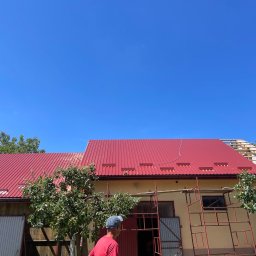 DACH-MAT Mateusz Russu - Czerwony blaszany dach na budynku gospodarczym w trakcie prac, widoczne rusztowanie i fragment drewnianej konstrukcji dachu w budowie, na pierwszym planie mężczyzna w czerwonej koszulce i czapce.