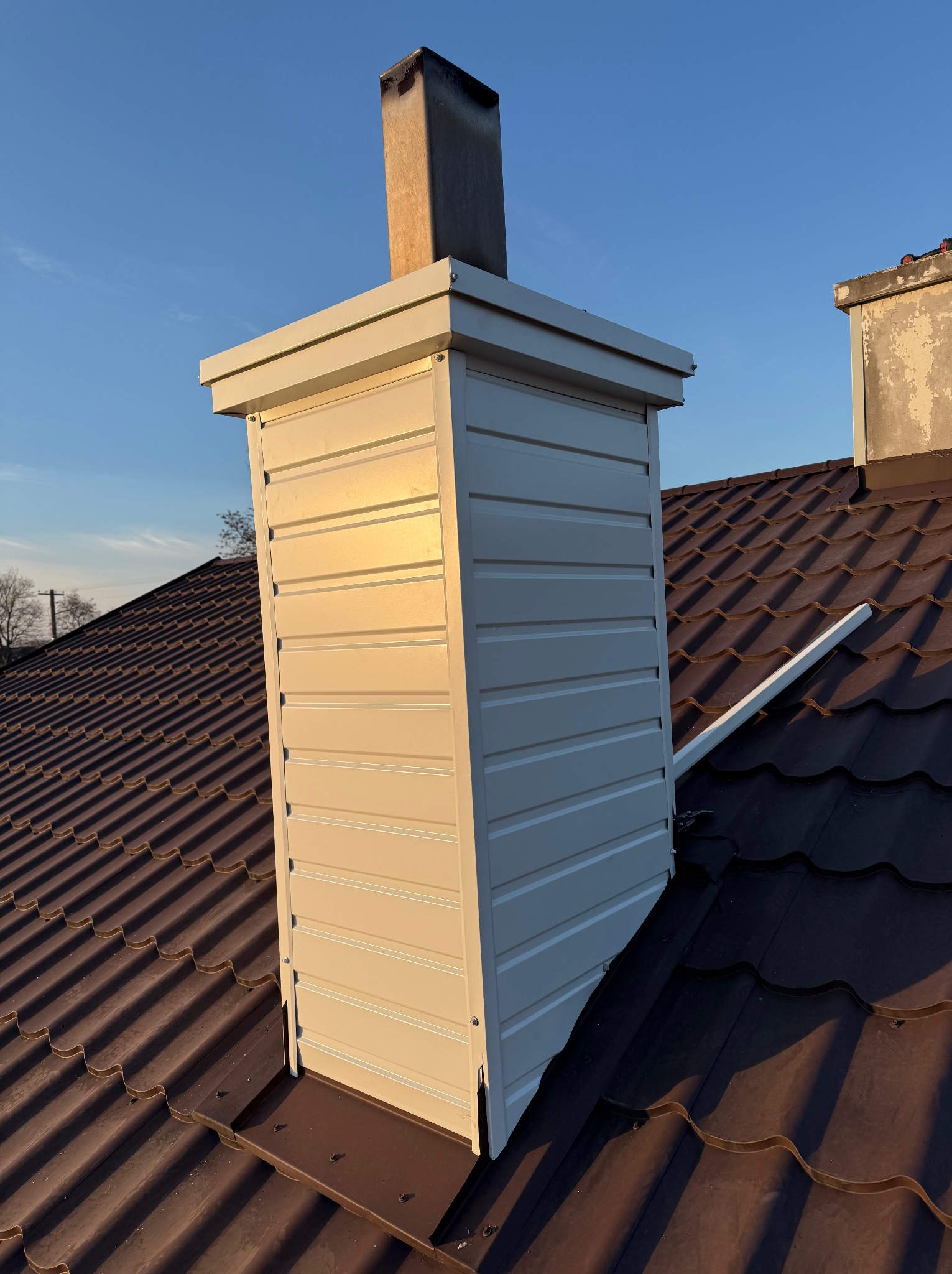 Obróbka blacharska komina z jasnej blachy na dachu z brązowej blachodachówki, widoczny fragment drugiego komina w tle, niebieskie niebo.