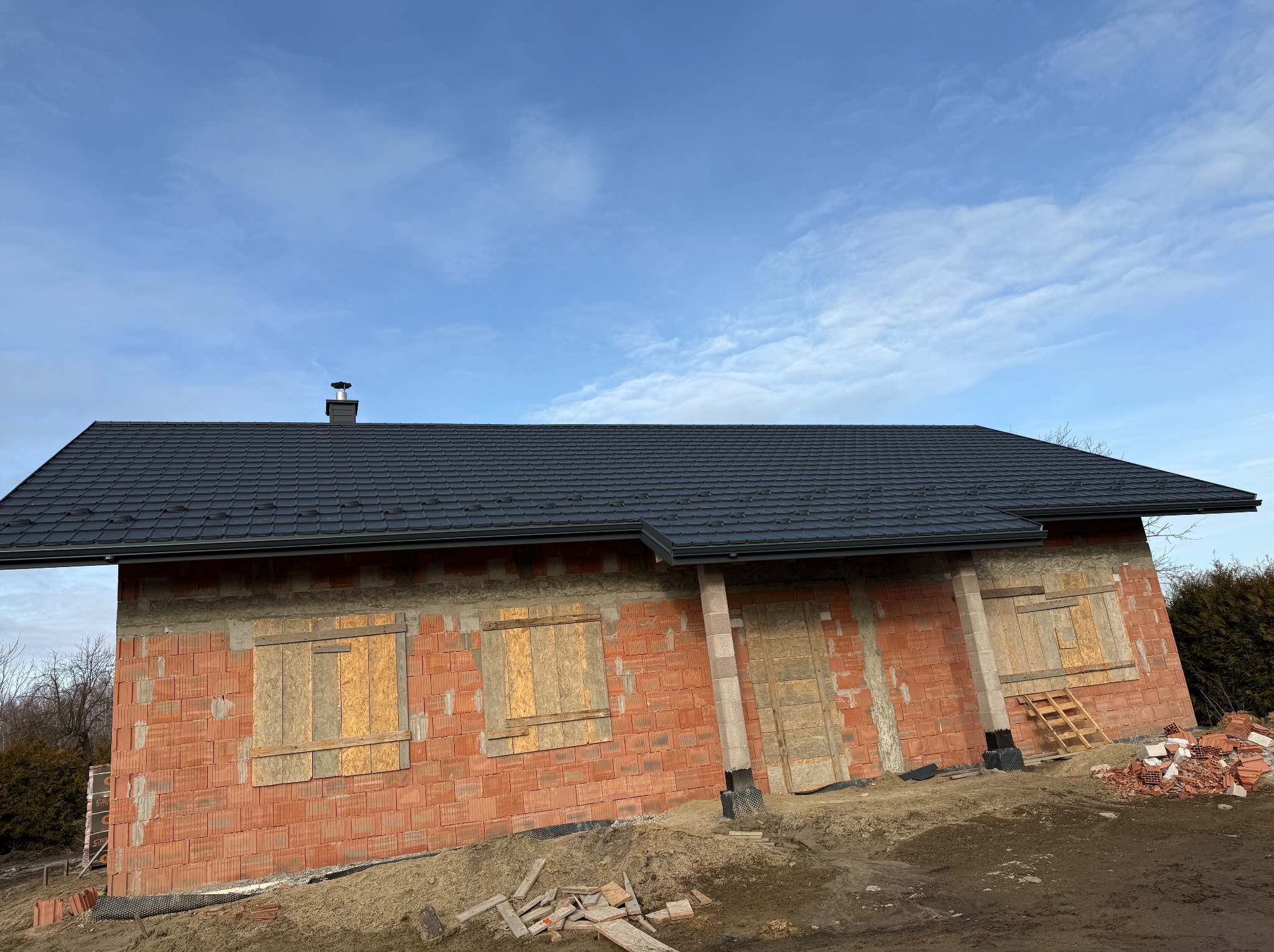 Dom w trakcie budowy z nowym, czarnym dachem, okna i drzwi zabite deskami, widoczne materiały budowlane i gruz.