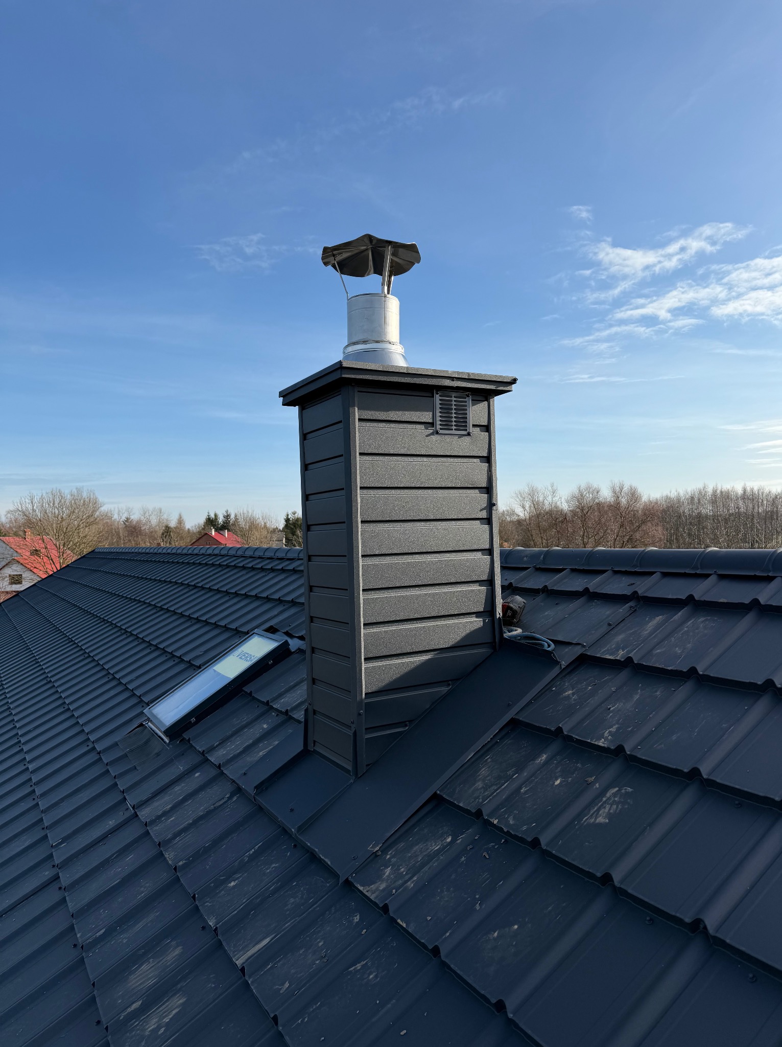 Nowy, ciemny dach z blachodachówki z oknem dachowym i kominem z metalową czapą, na tle błękitnego nieba.