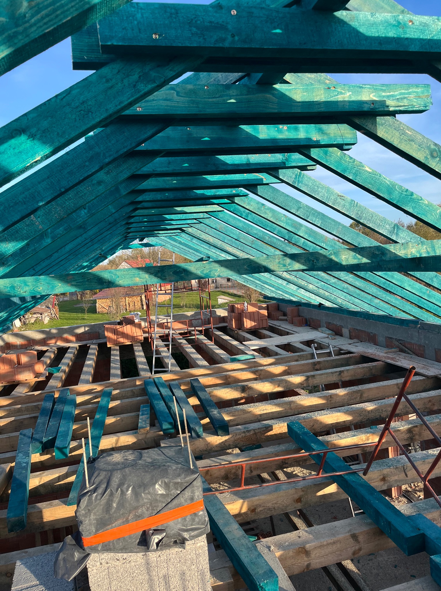 Konstrukcja drewniana dachu, impregnowana na zielono, widok z wnętrza budynku, przygotowana pod dalsze prace dekarskie, z widocznymi cegłami i elementami rusztowania w tle.