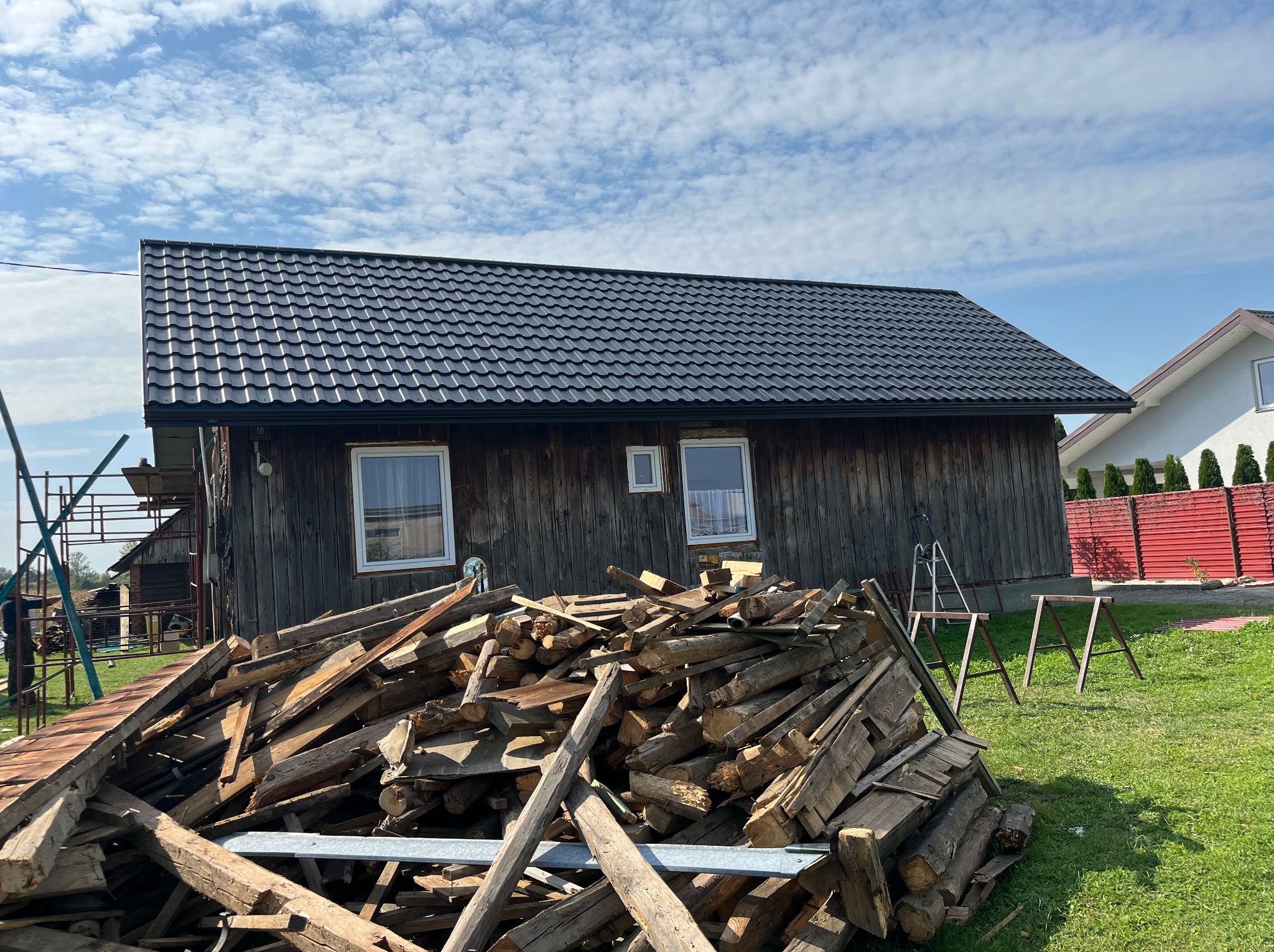Drewniany budynek z nowym, czarnym dachem z blachodachówki, na pierwszym planie sterta drewna i rusztowanie, w tle błękitne niebo z chmurami.