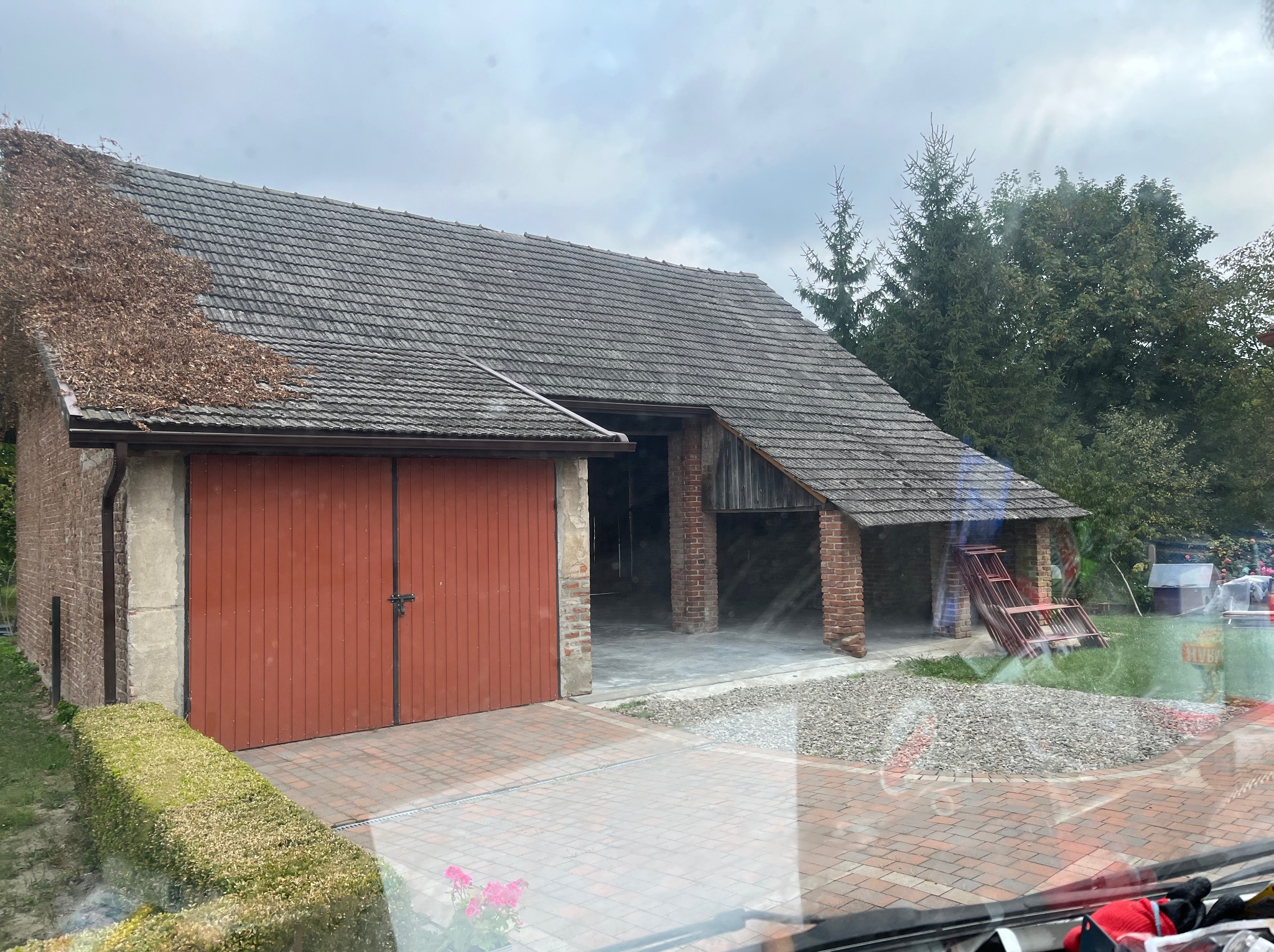 Widok na budynek gospodarczy z dwuspadowym dachem pokrytym szarą dachówką, częściowo porośniętym suchymi pnączami, z czerwonymi wrotami garażowymi i otwartą wiatą z ceglanymi filarami.