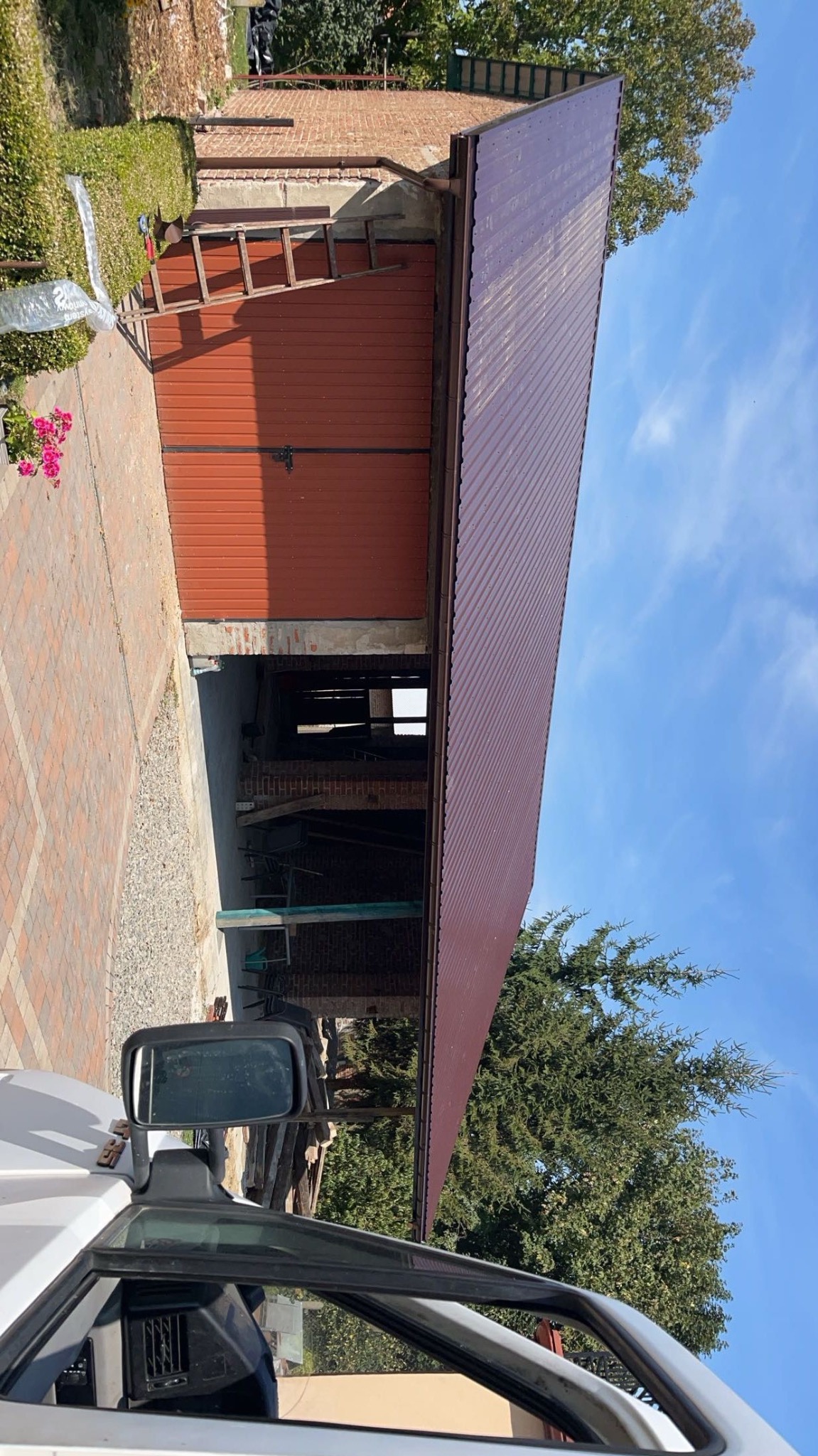 Nowy blaszany dach w kolorze bordowym na budynku gospodarczym w Wietlinie Pierwszym, widok zza szyby samochodu, z boku widoczna drabina oparta o budynek.