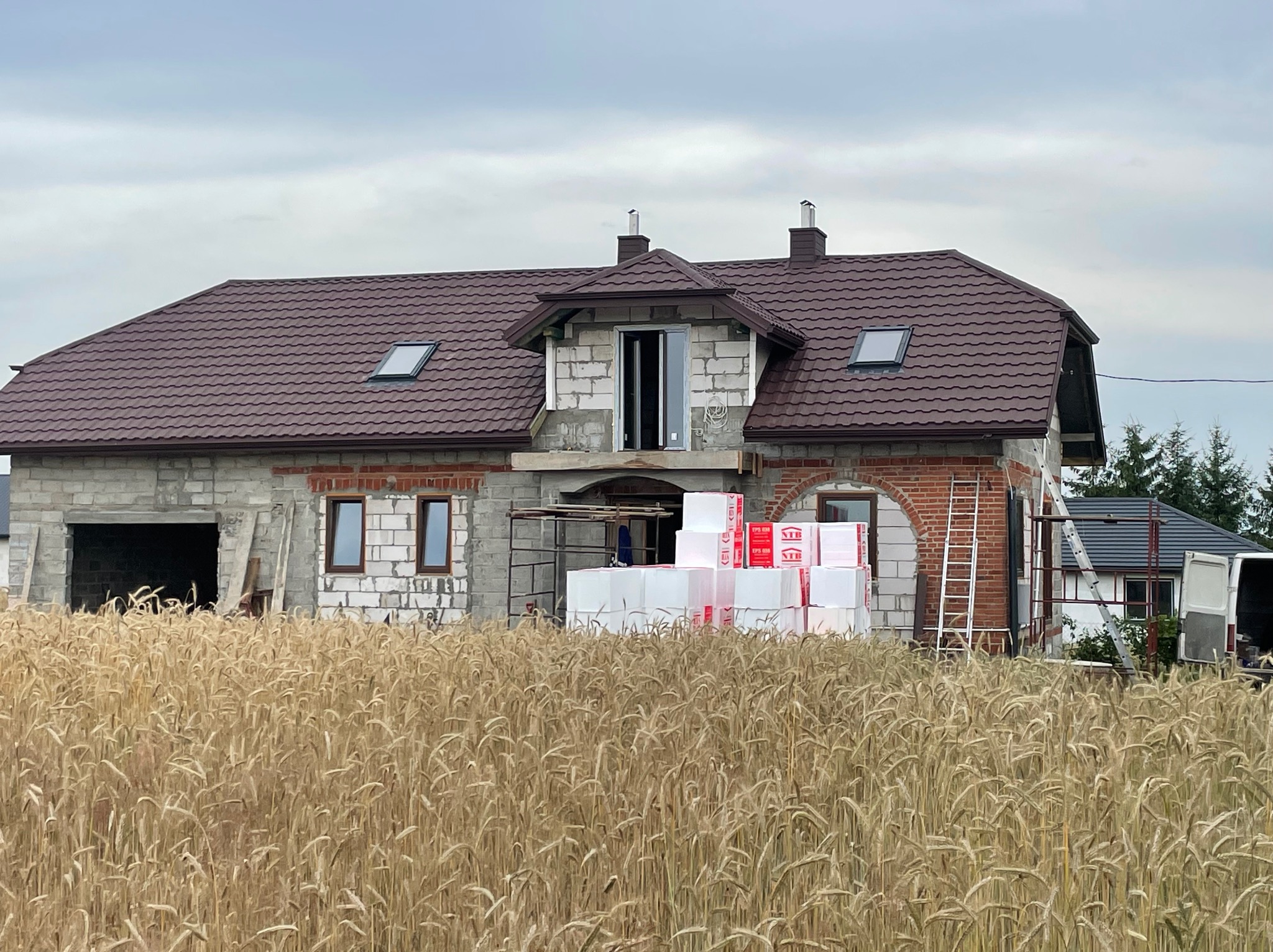 Dom w trakcie budowy z brązowym dachem, widoczne okna dachowe, kominy, rusztowania, materiały izolacyjne i pole zboża na pierwszym planie.